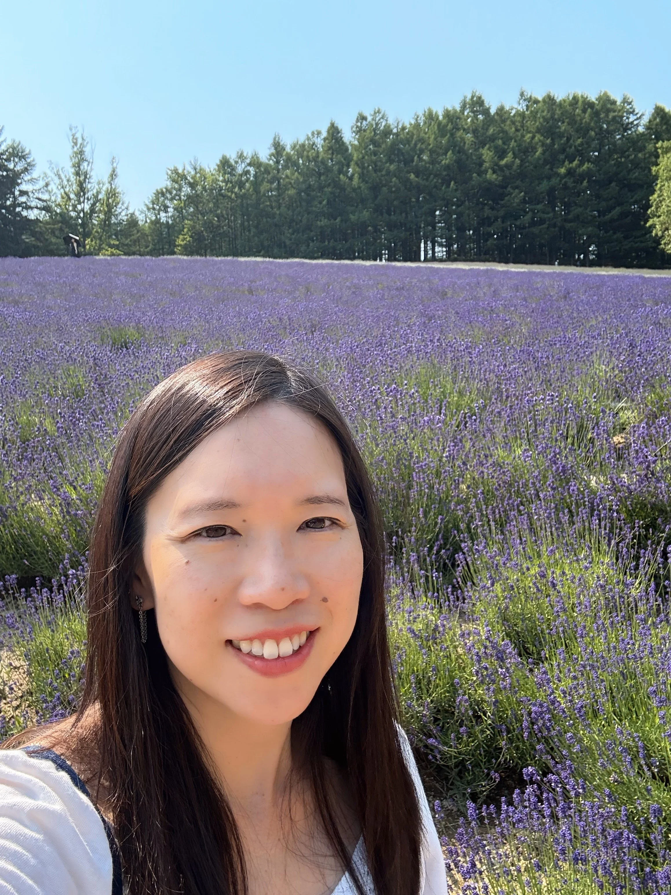 A woman taking a selfie in a lavender field on a sunny day with a clear blue sky and trees in the background.