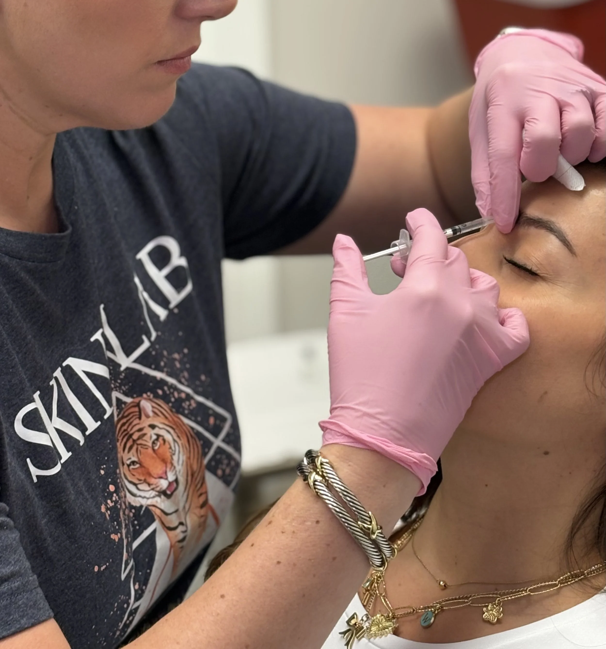 A woman receives a cosmetic injection in her forehead from a professional wearing pink gloves. The professional is holding a syringe, and the woman has her eyes closed. The woman is wearing jewelry, including necklaces and bracelets, and a dark T-shirt with a tiger and the word 'SKINLAB' printed on it.