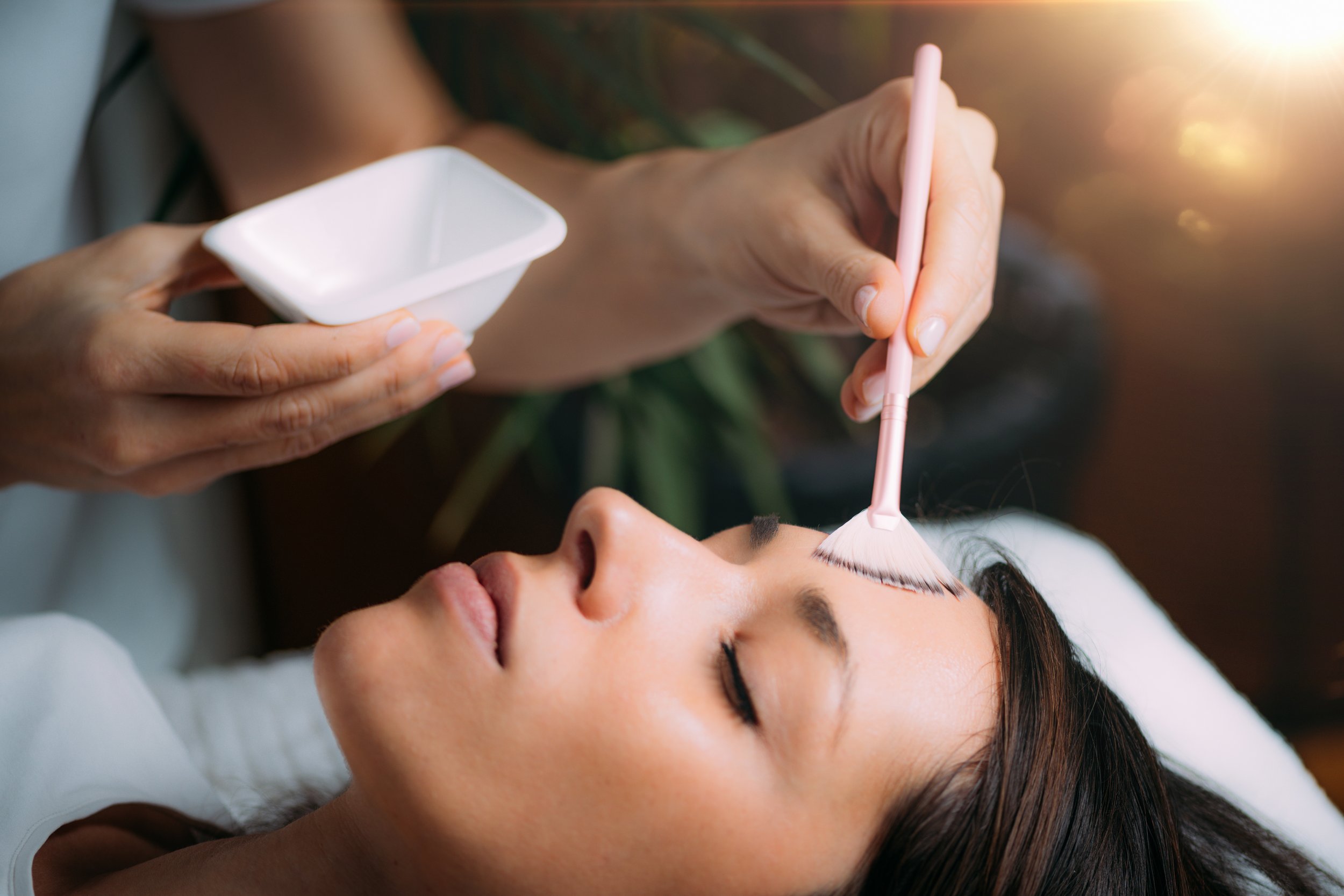 A woman receiving a cosmetic eyelash treatment with a brush, while another person holds a small bowl.