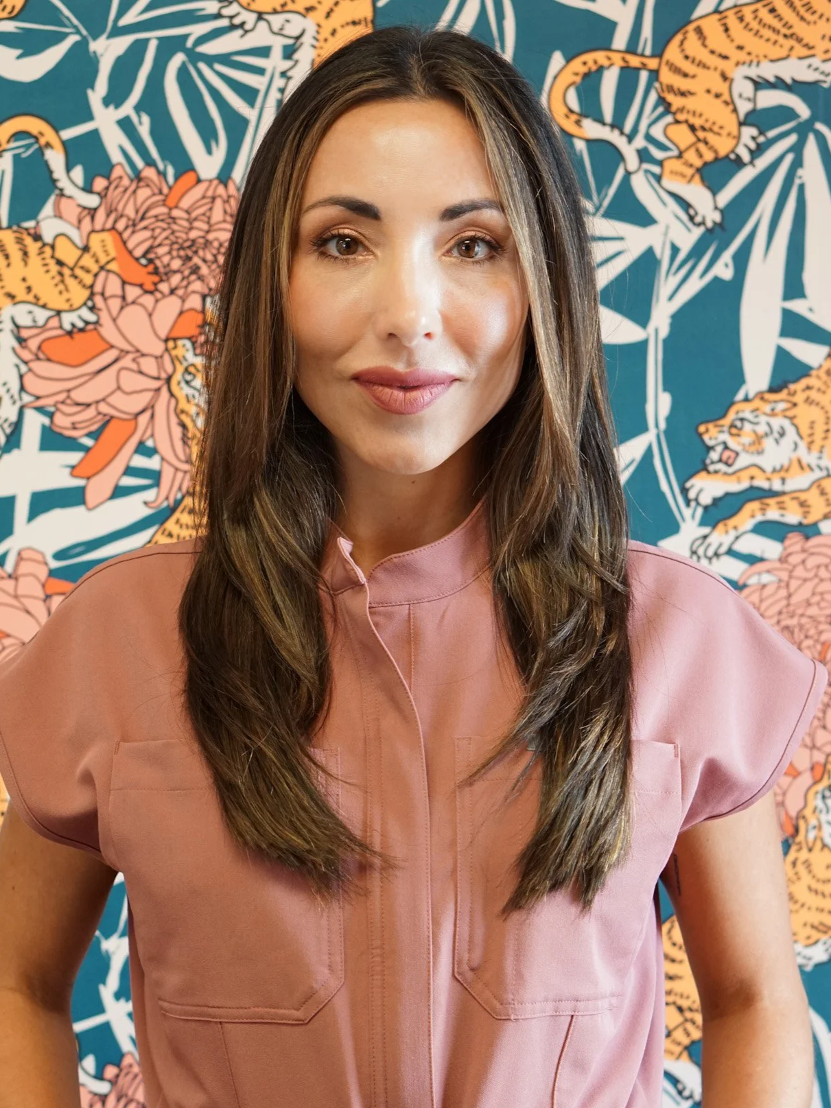 A woman with long brown hair and light makeup, wearing a pink blouse, stands in front of a colorful jungle-themed mural with tigers and flowers.