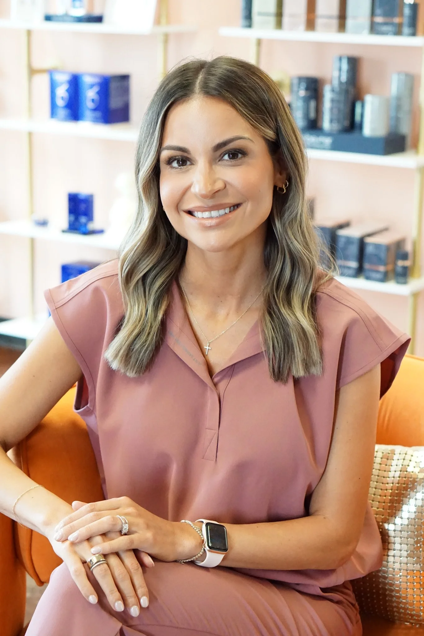 A woman with wavy, shoulder-length hair smiling, sitting on an orange chair, wearing a pink short-sleeve top, jewelry, and a smartwatch, with a bookshelf in the background.