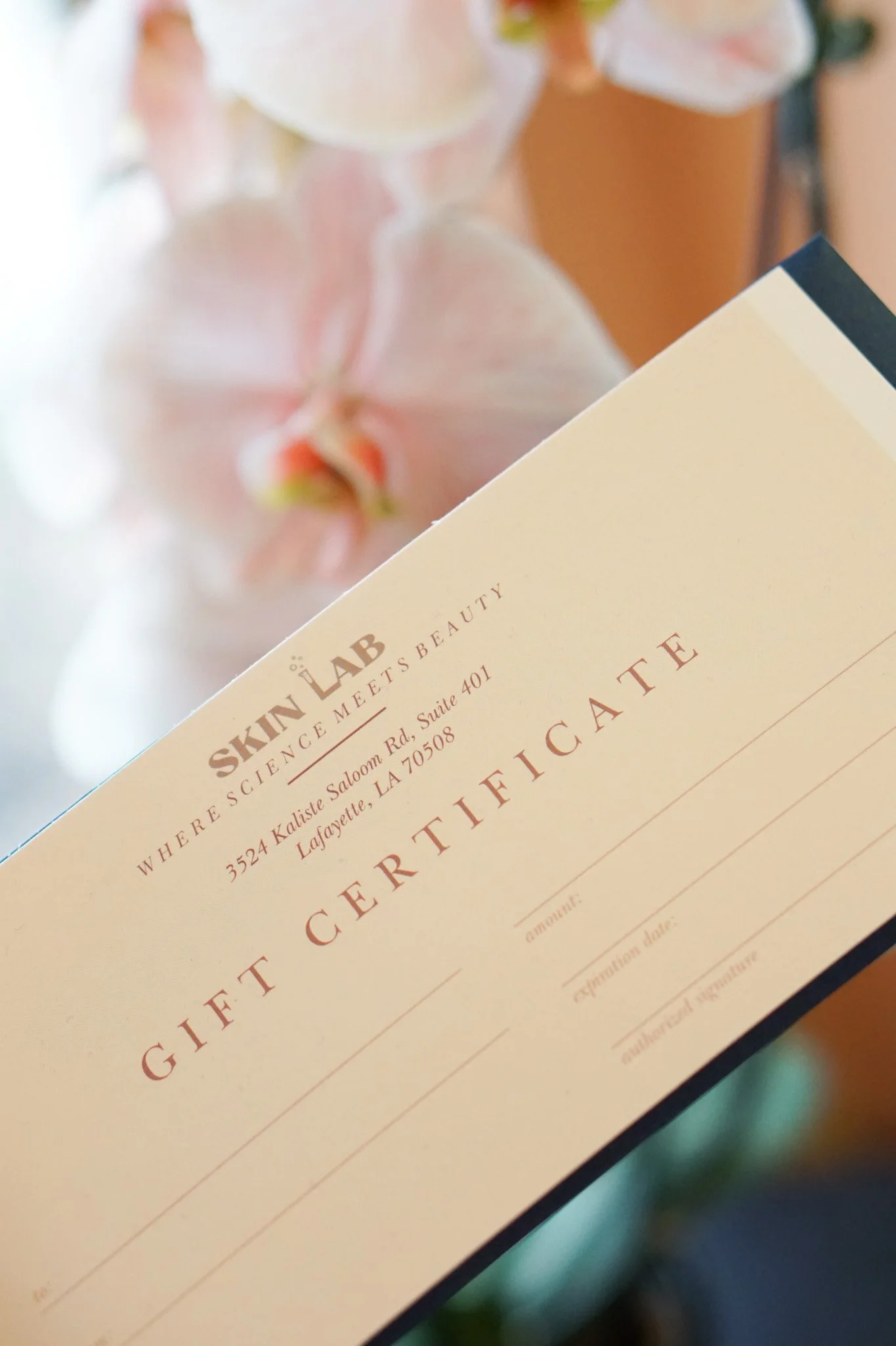 A gift certificate from Skin Lab, with the address 3524 Kaliste Saloom Rd., Suite 401, Lafayette, LA 70508, placed at an angle in front of some blurred pink and white orchids.
