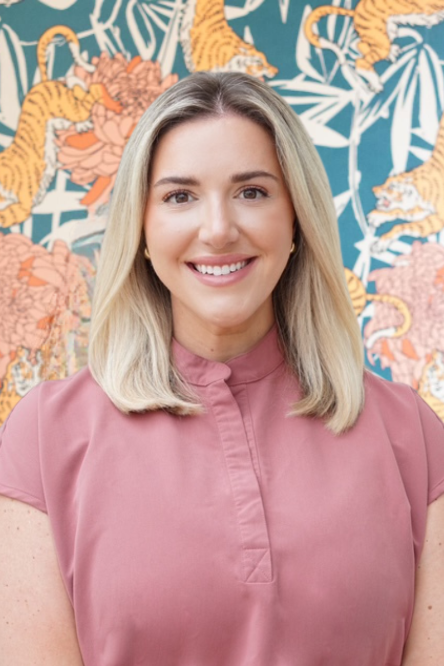 A woman with blonde hair smiling, wearing a pink top, standing in front of a colorful tiger and floral patterned background.