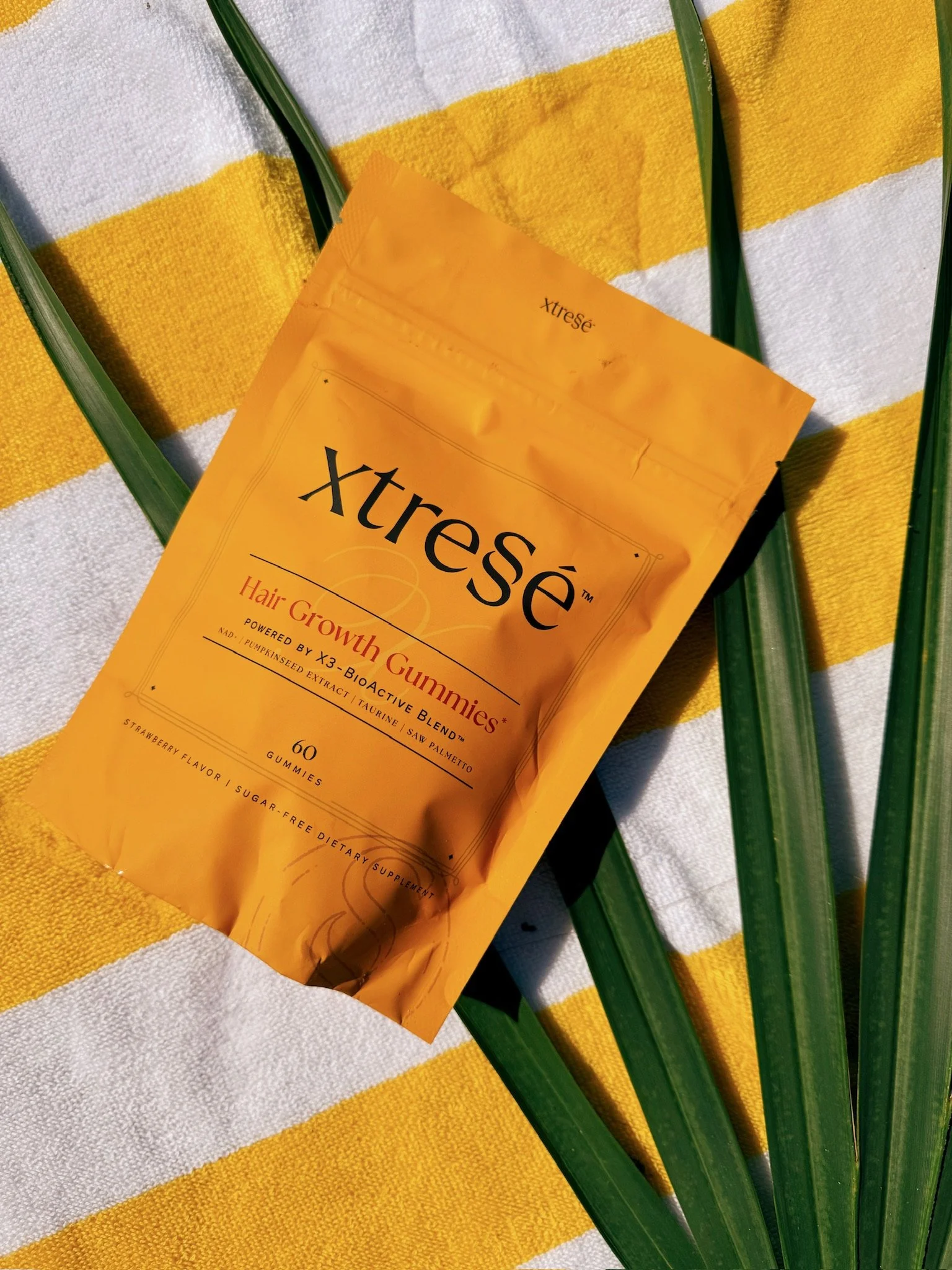 A yellow packet of Xtress Hair Growth Gummies resting on a yellow and white striped towel with green palm fronds nearby.