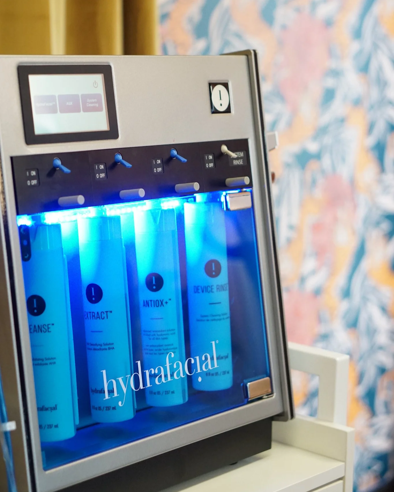 Hydrafacial machine with bottles labeled 'HYDRAFACIAL', illuminated by blue light, set against a background with a floral pattern wallpaper.