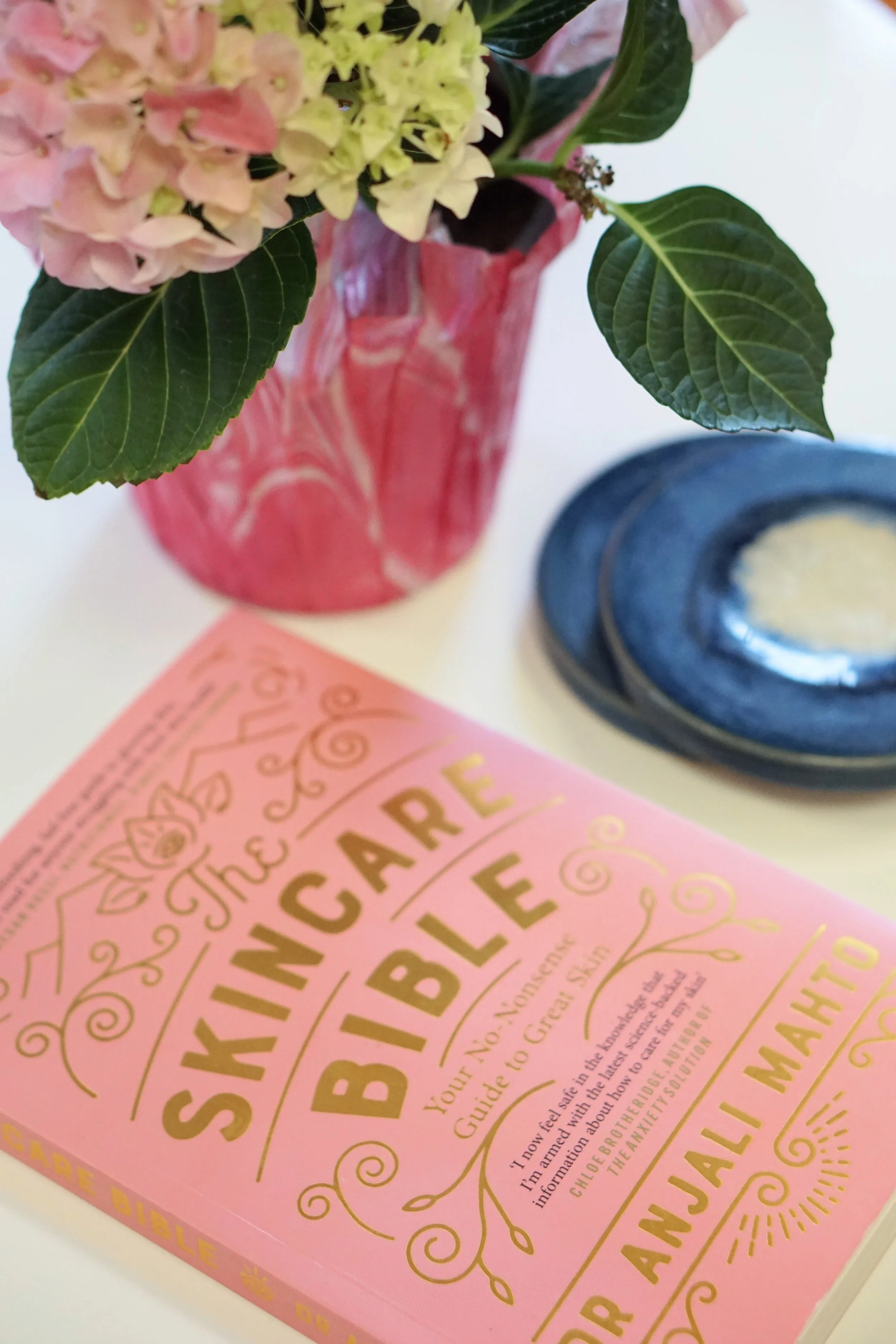 A pink book titled 'The Skeincare Bible' lies on a white surface, with a red vase holding a hydrangea flower in pink and green hues nearby. Part of a blue dish with a light-colored substance is also visible.