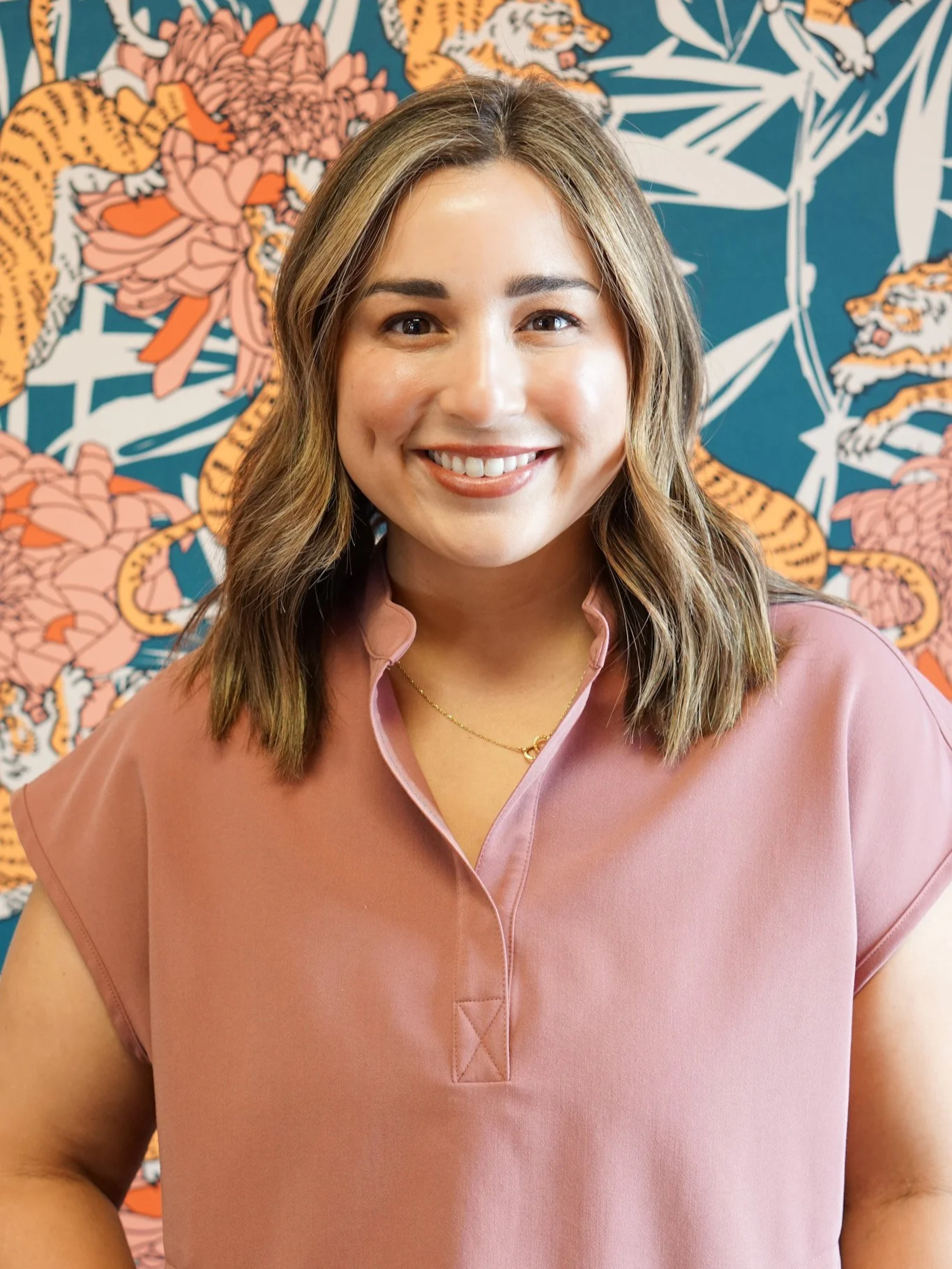 A smiling woman with shoulder-length brown hair wearing a pink shirt standing in front of a colorful mural with orange tigers and flowers.