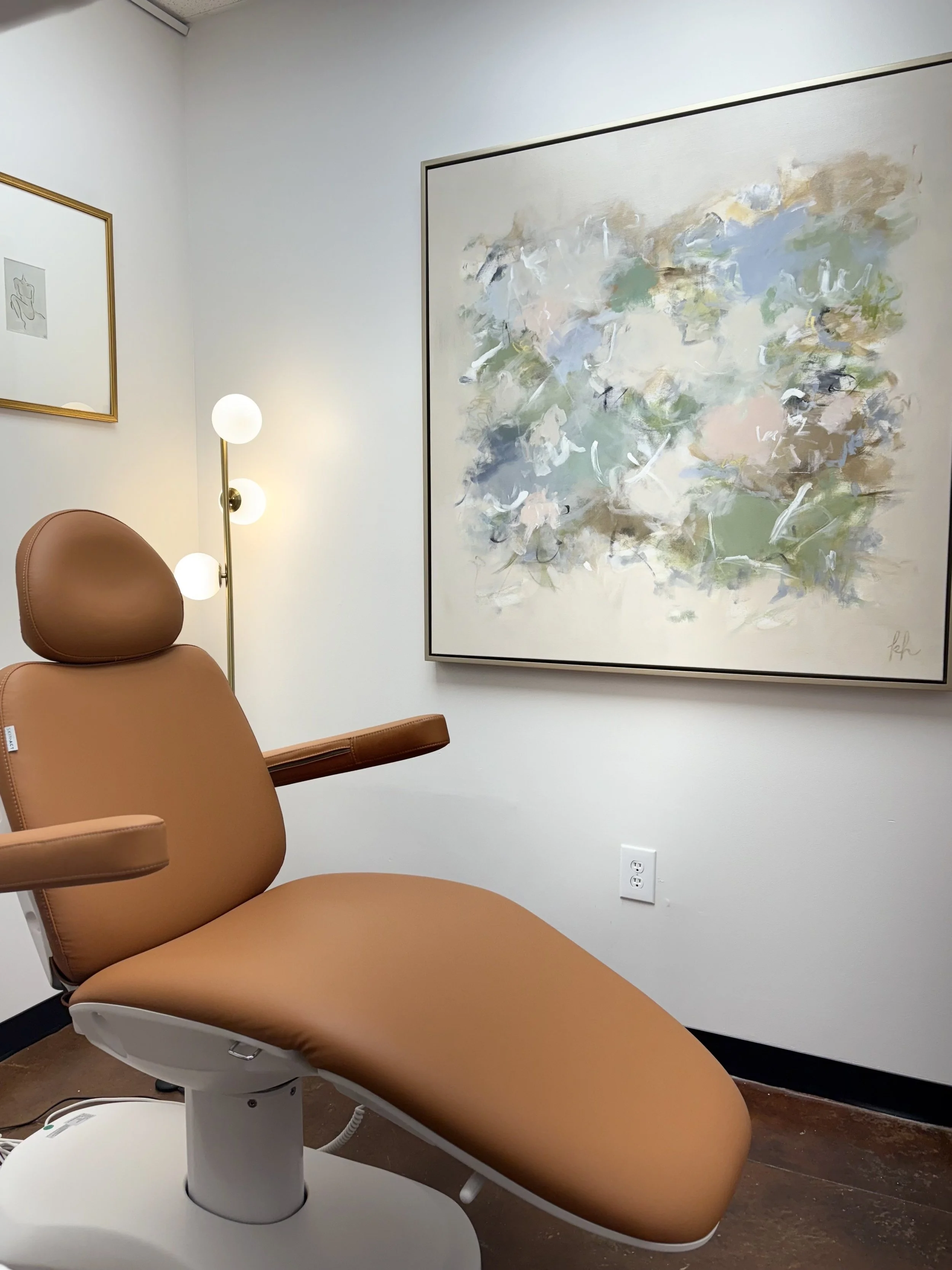 An examination room with a brown medical chair, a floor lamp with three white globe lights, and abstract artwork on the wall.