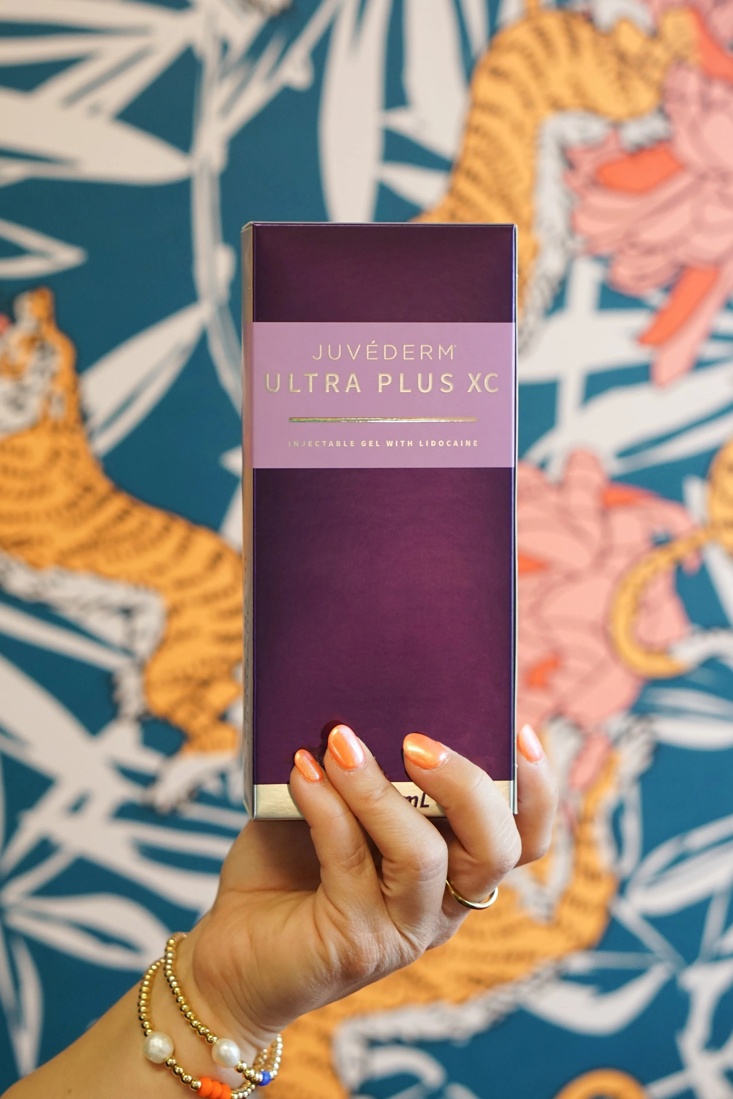 Hand holding a purple box of Juvederm Ultra Plus XC, a dermal filler product, with a colorful tiger and floral wallpaper in the background.