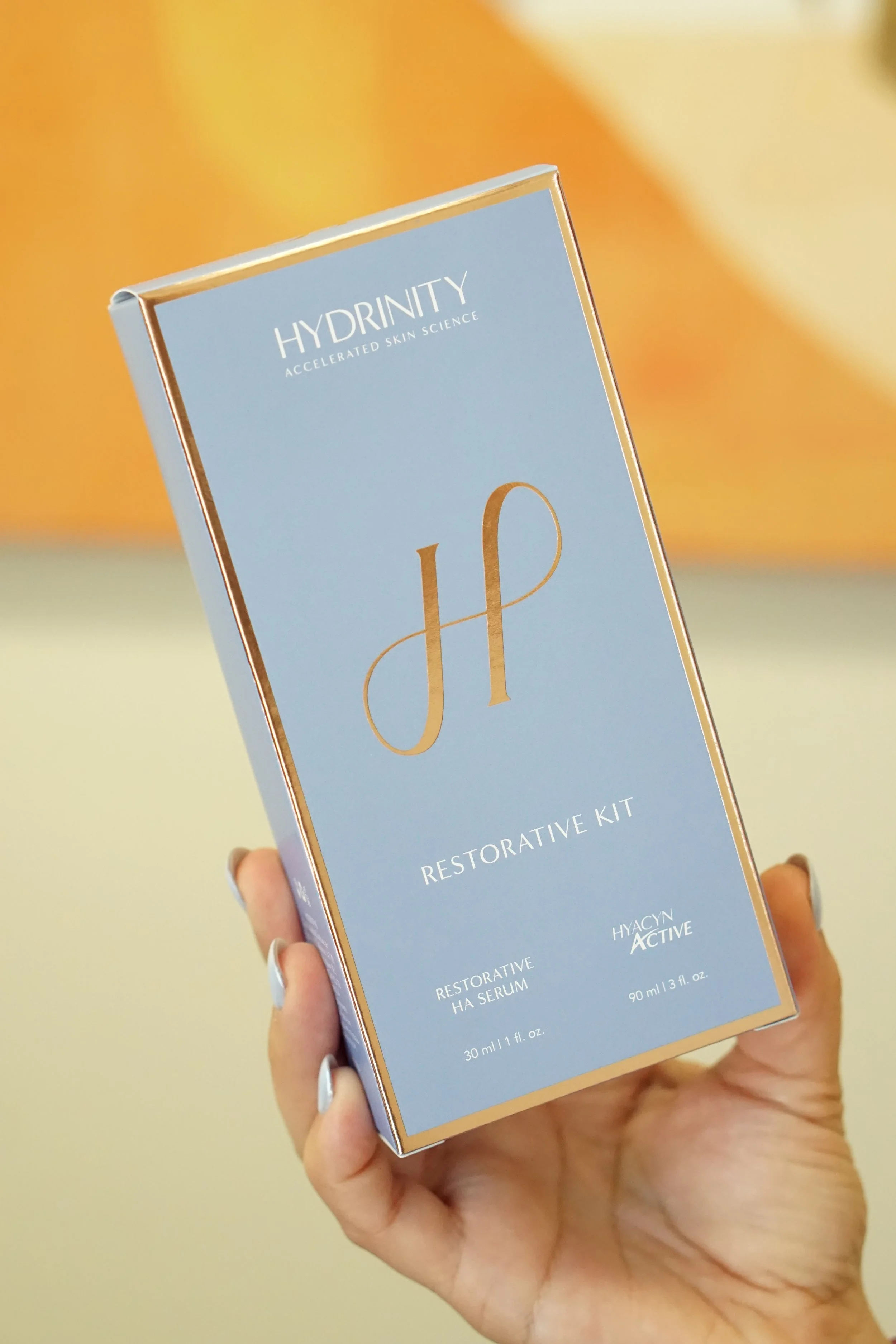 A hand holding a blue box with gold accents and text that reads 'Hydrinity Restorative Kit' and mentions serum and active ingredients.