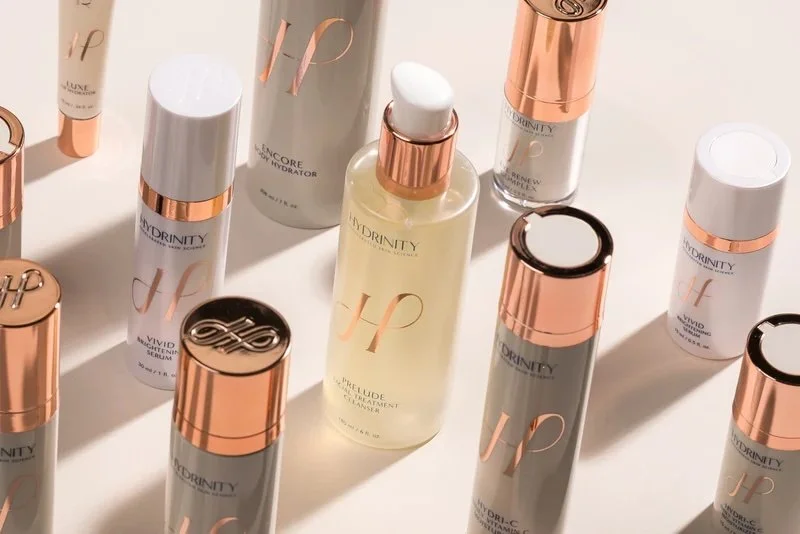Assorted skincare products from Hydrdinity, including bottles and tubes with rose gold and white accents, arranged on a white surface.