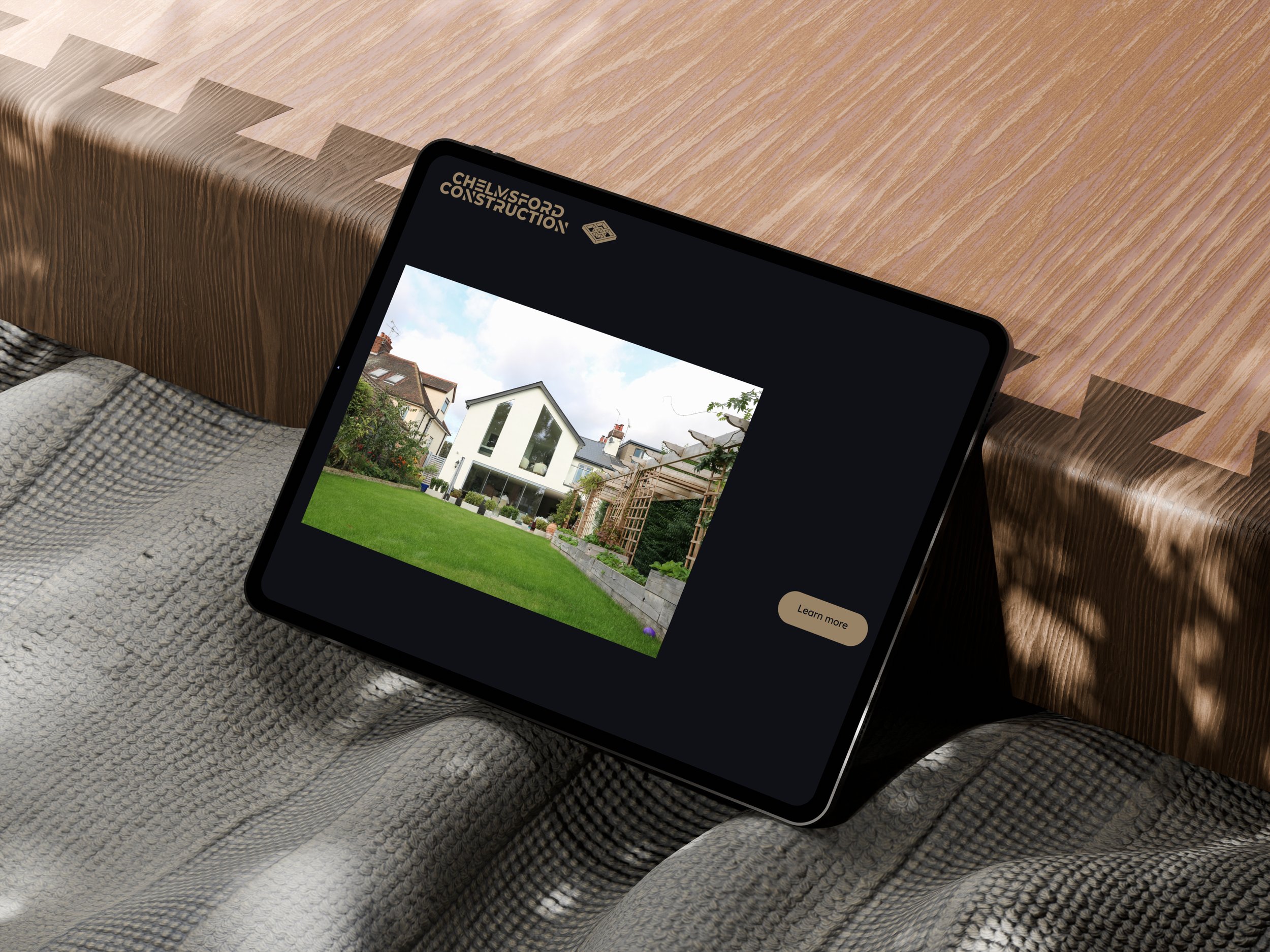 Tablet displaying a modern house in a yard with green grass, trees, and construction scaffolding on the side, placed on a textured blanket on a bed next to a wooden headboard.