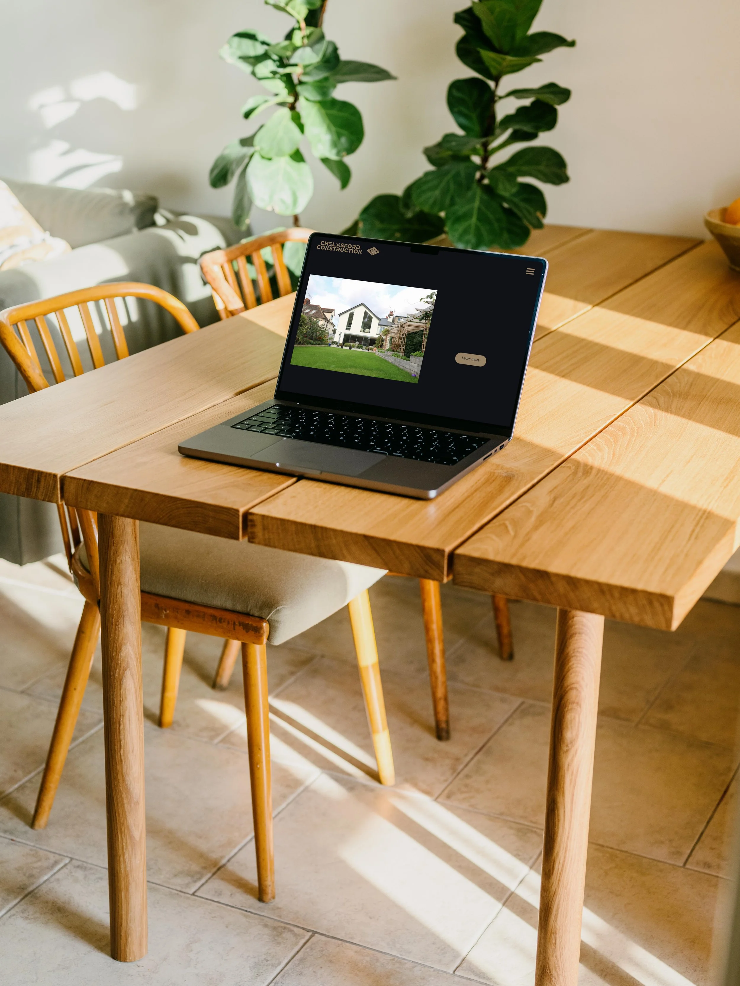 A wooden dining table with a laptop displaying a house design. There are wooden chairs around the table, and a plant is visible in the background.