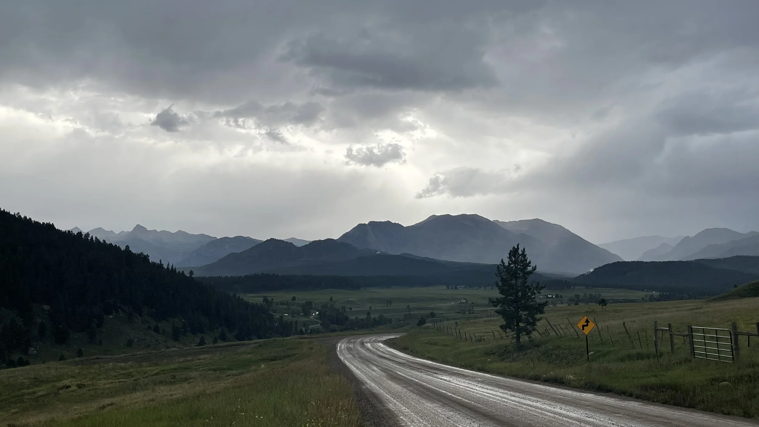 A winding dirt road through a green valley with mountains in the background under a cloudy sky.