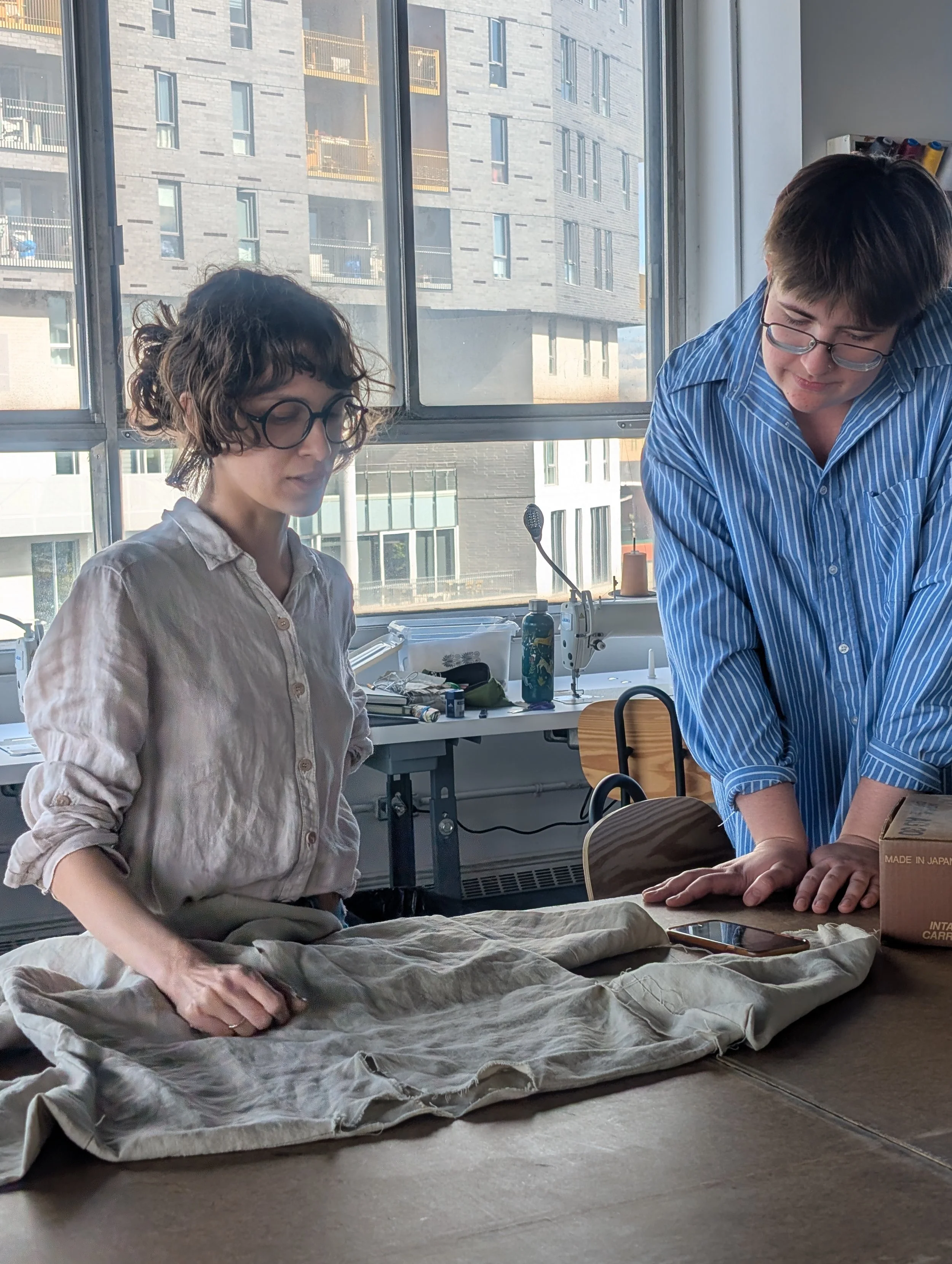 Deux individus se consultent autour d'une pièce textile, dans un atelier éclairé par une grande fenêtre.