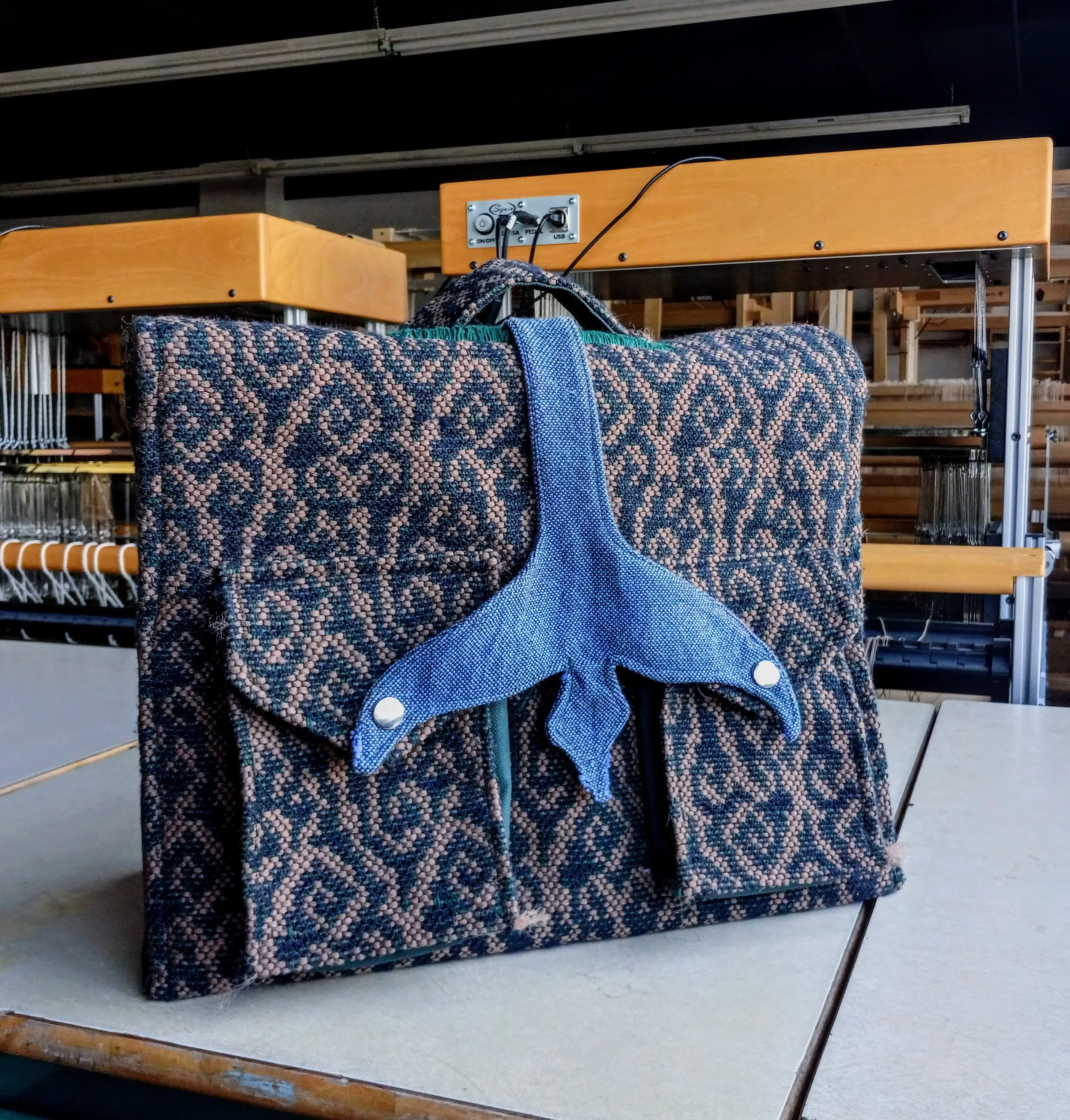 Le sac corbeau tissé à la main, dans des motifs de rinceaux verts et bruns, avec une sangle pervenche en forme d'oiseau, et des éléments métalliques. Il y a un métier à tisser derrière.