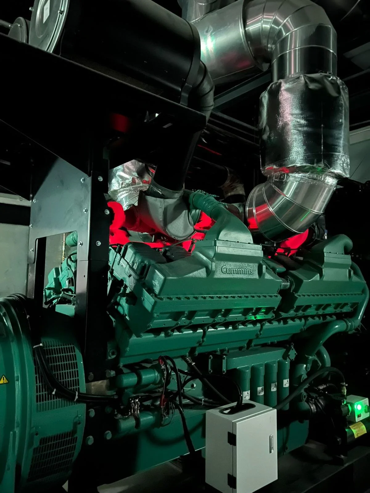 Industrial engine with visible pipes and components, including a green Cummins engine and metallic ductwork, in a mechanical room.