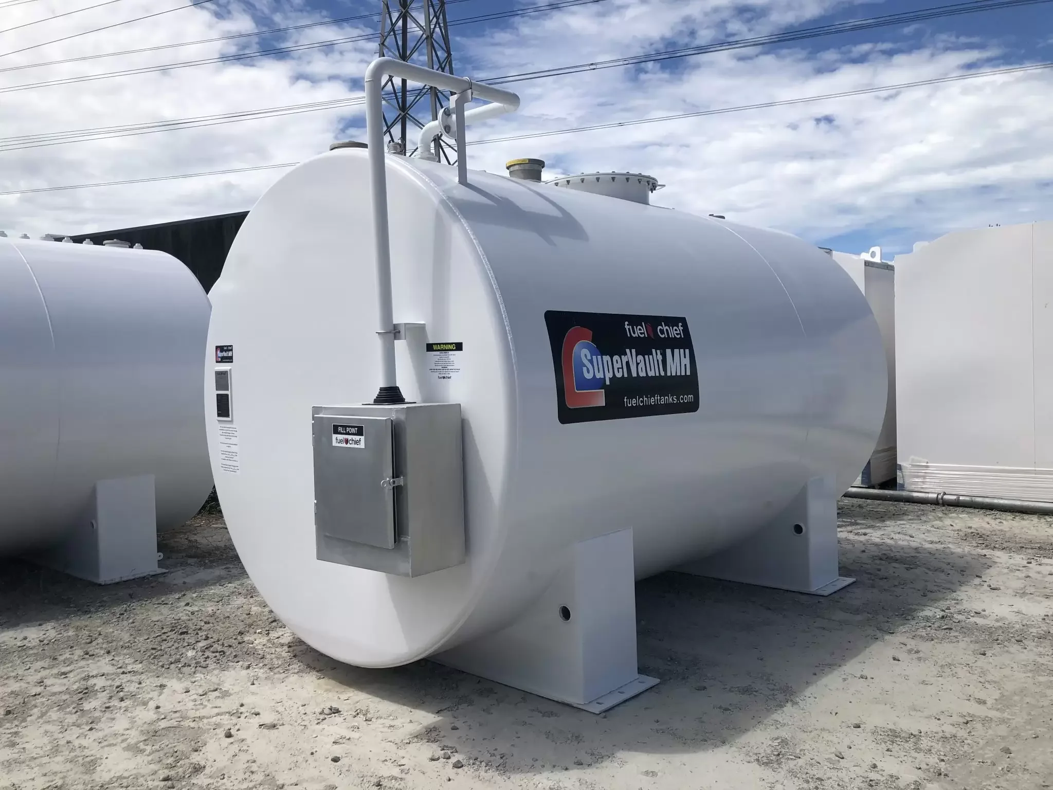 Large white fuel tank labeled 'SuperVault MH' with a smaller gray control box attached, outdoors on a gravel surface with power lines and a blue sky with clouds in the background.