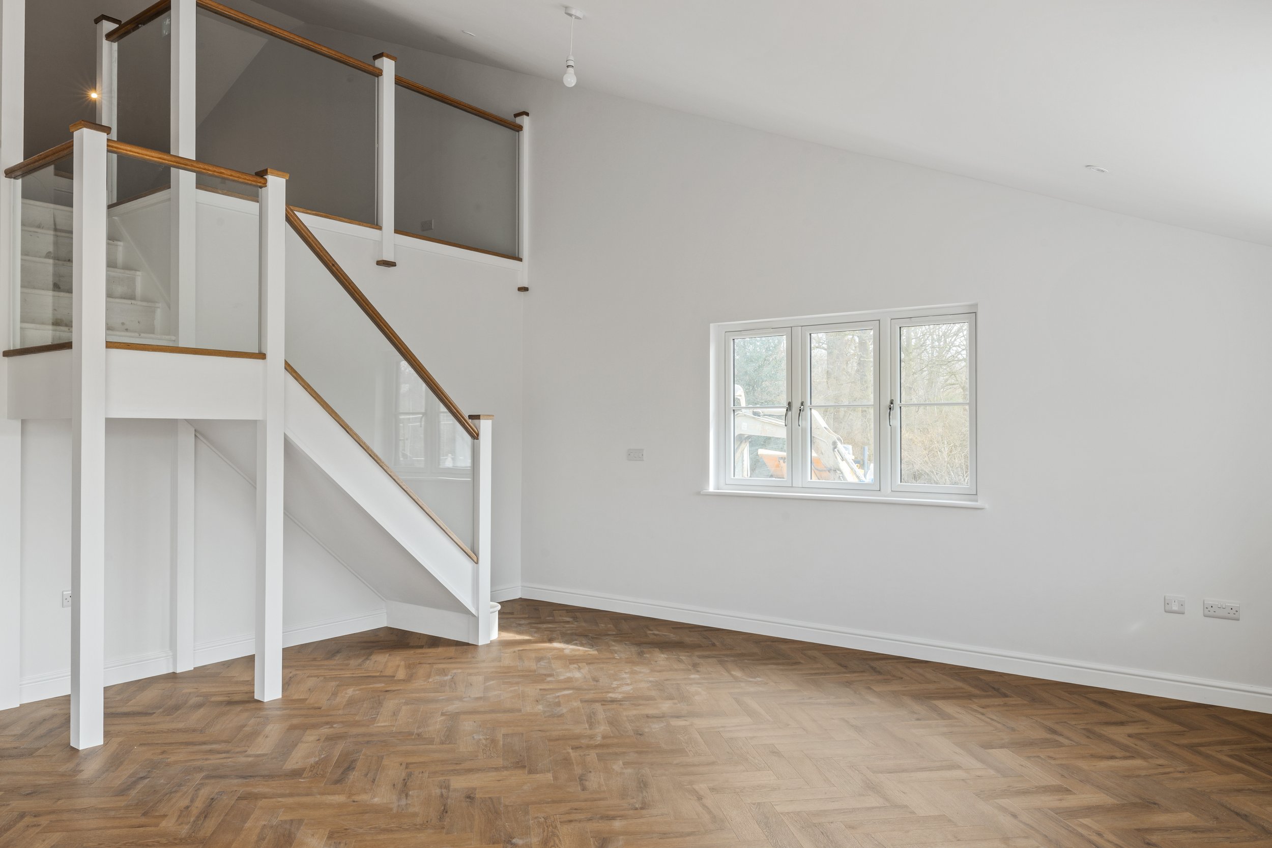 Empty living room with wooden floors, white walls, window, and staircase with wooden handrails.