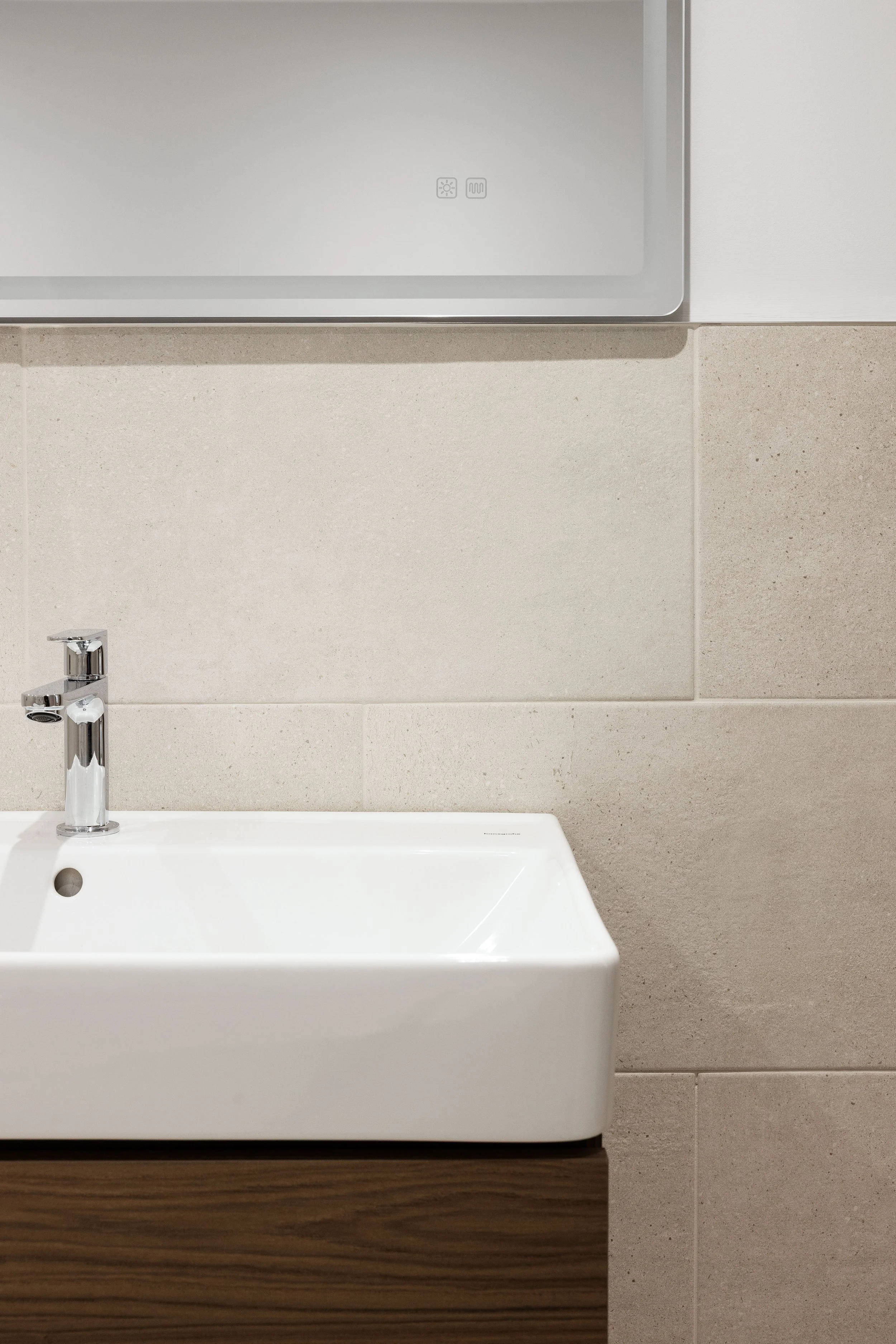 Bathroom sink with faucet and mirror above, beige tiled wall, wooden cabinet below.