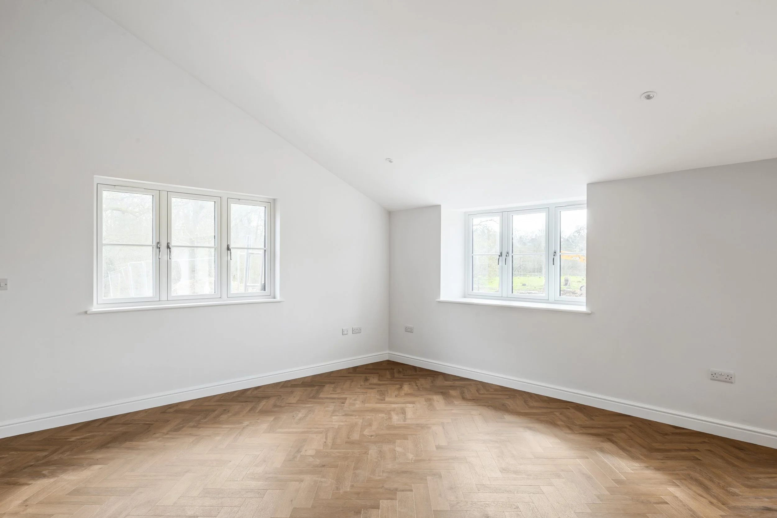 Empty white room with hardwood floors and three windows showing greenery outside.