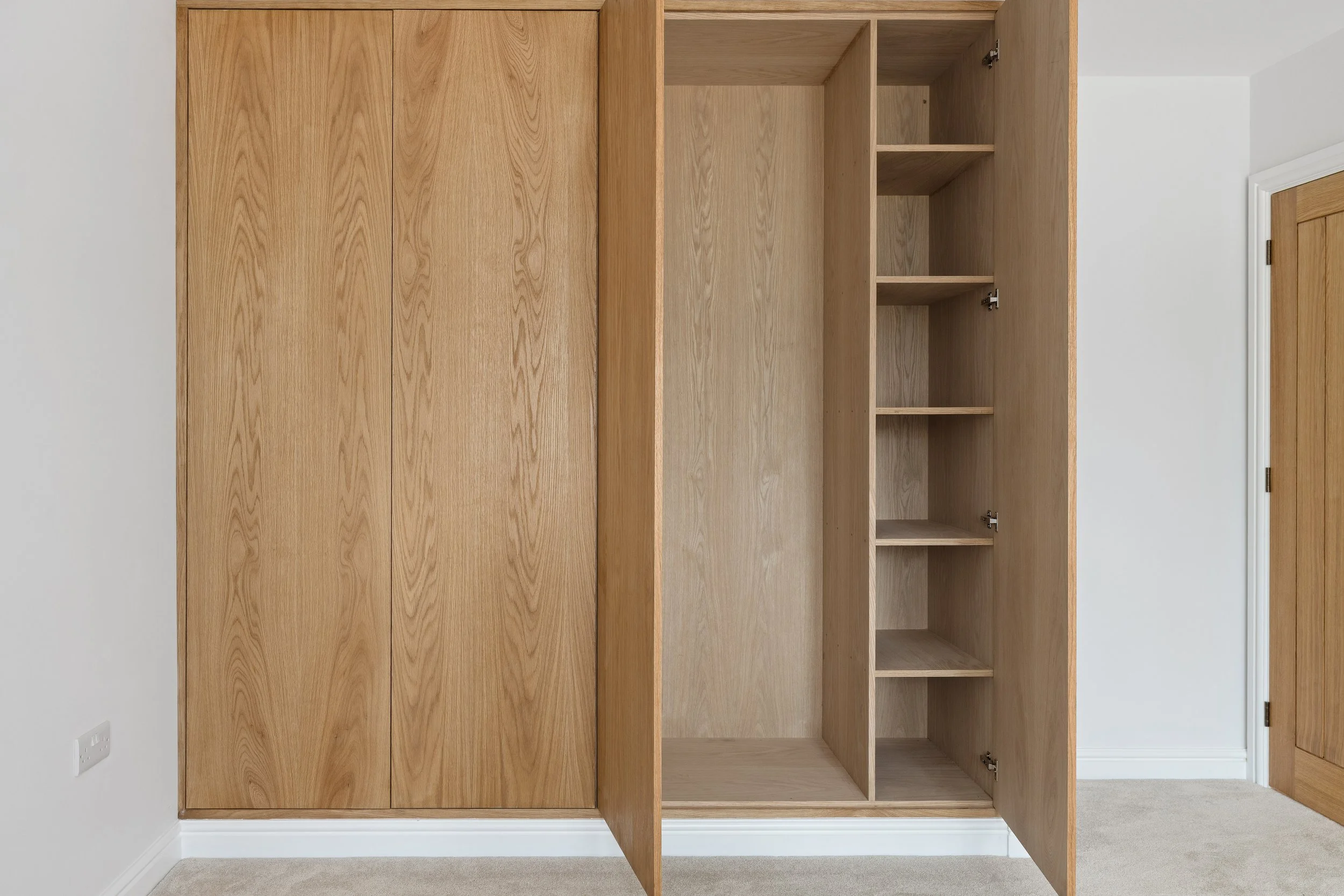 Empty wooden wardrobe with open doors revealing shelves inside, located in a room with white walls and a beige carpet.