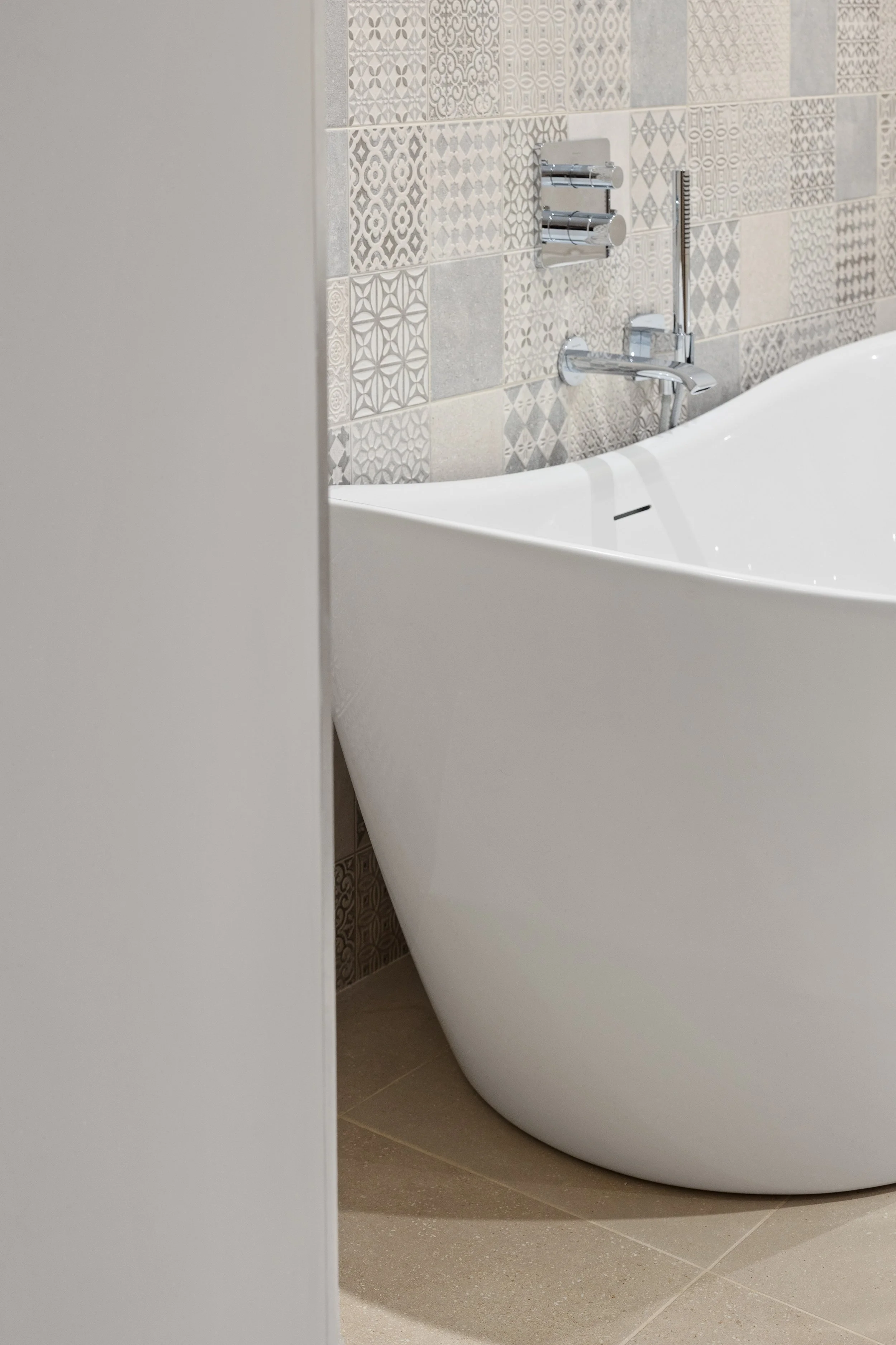 Part of a modern bathroom with a white freestanding bathtub, decorative tiled wall, and chrome fixtures.