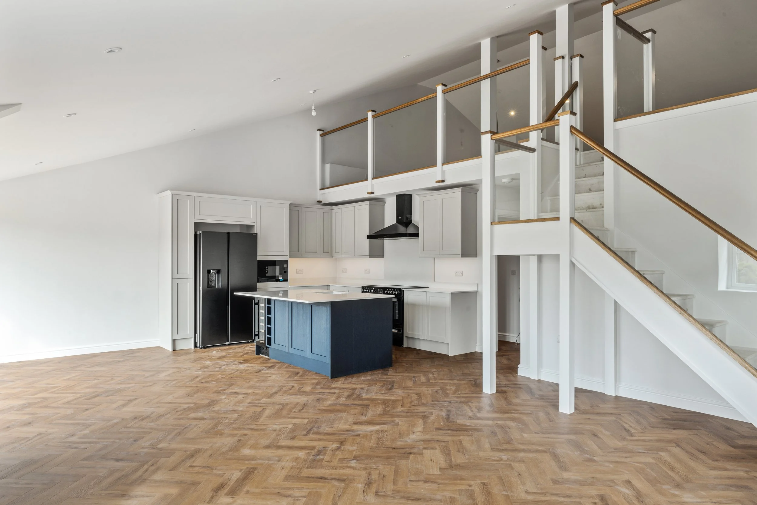 Empty modern kitchen with white cabinets, black refrigerator, stove, and range hood, open floor plan with wooden flooring, staircase with wooden handrails, and an upper-level balcony overlooking the space.