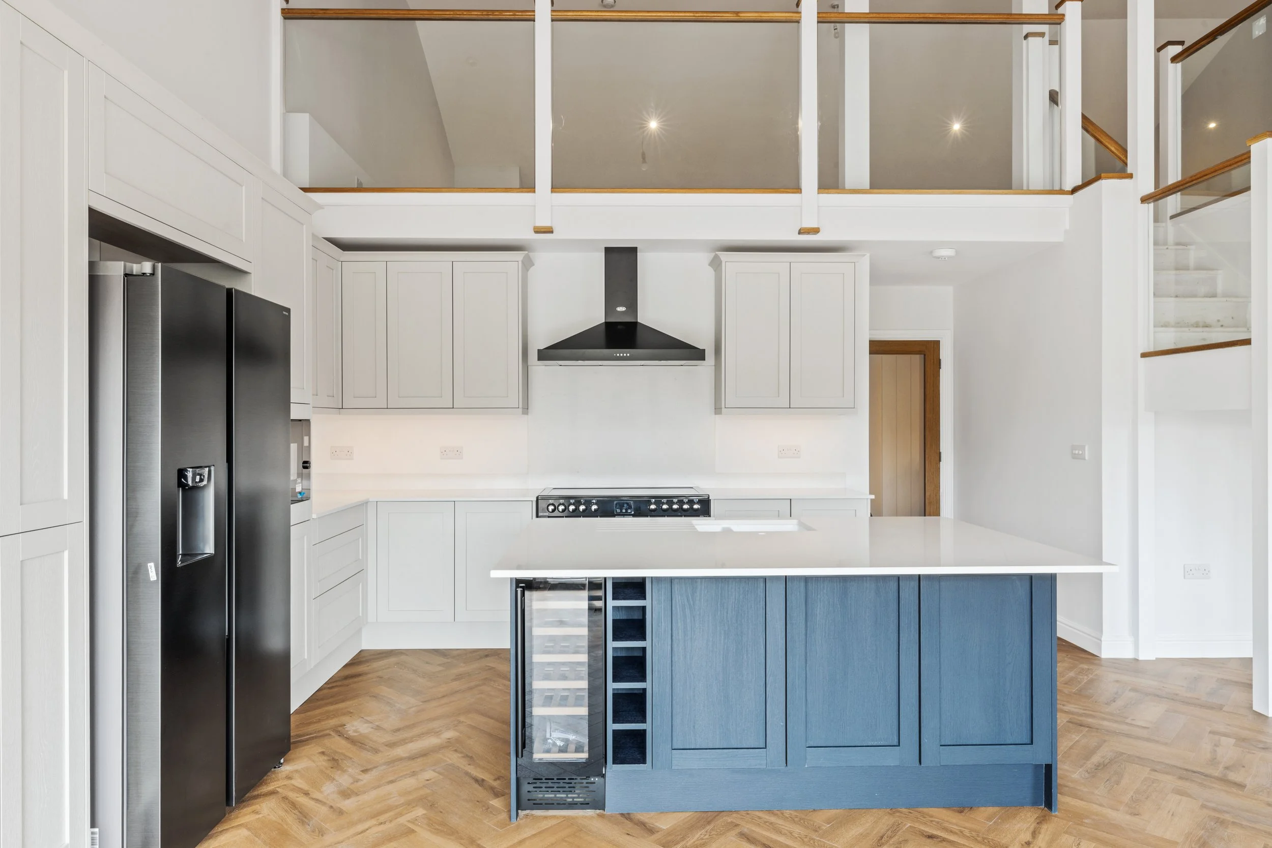 Modern kitchen with white cabinets, black refrigerator, blue kitchen island with wine rack and mini fridge, stainless steel stove, black range hood, wooden flooring, staircase, and an open upper level with railing.