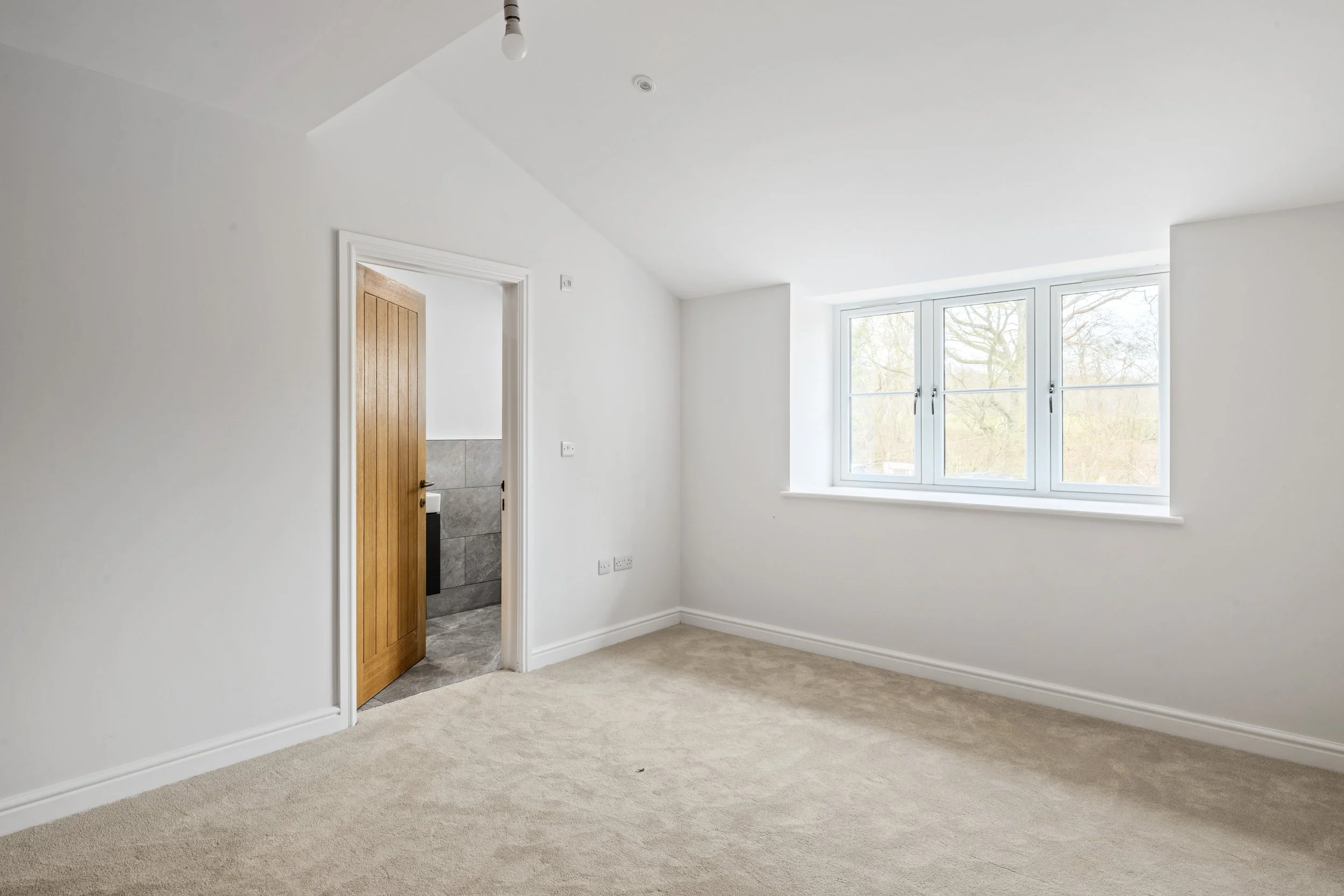Empty room with white walls, beige carpet, a window with four panes letting in natural light, and a wooden door leading to a bathroom with gray tiled walls.