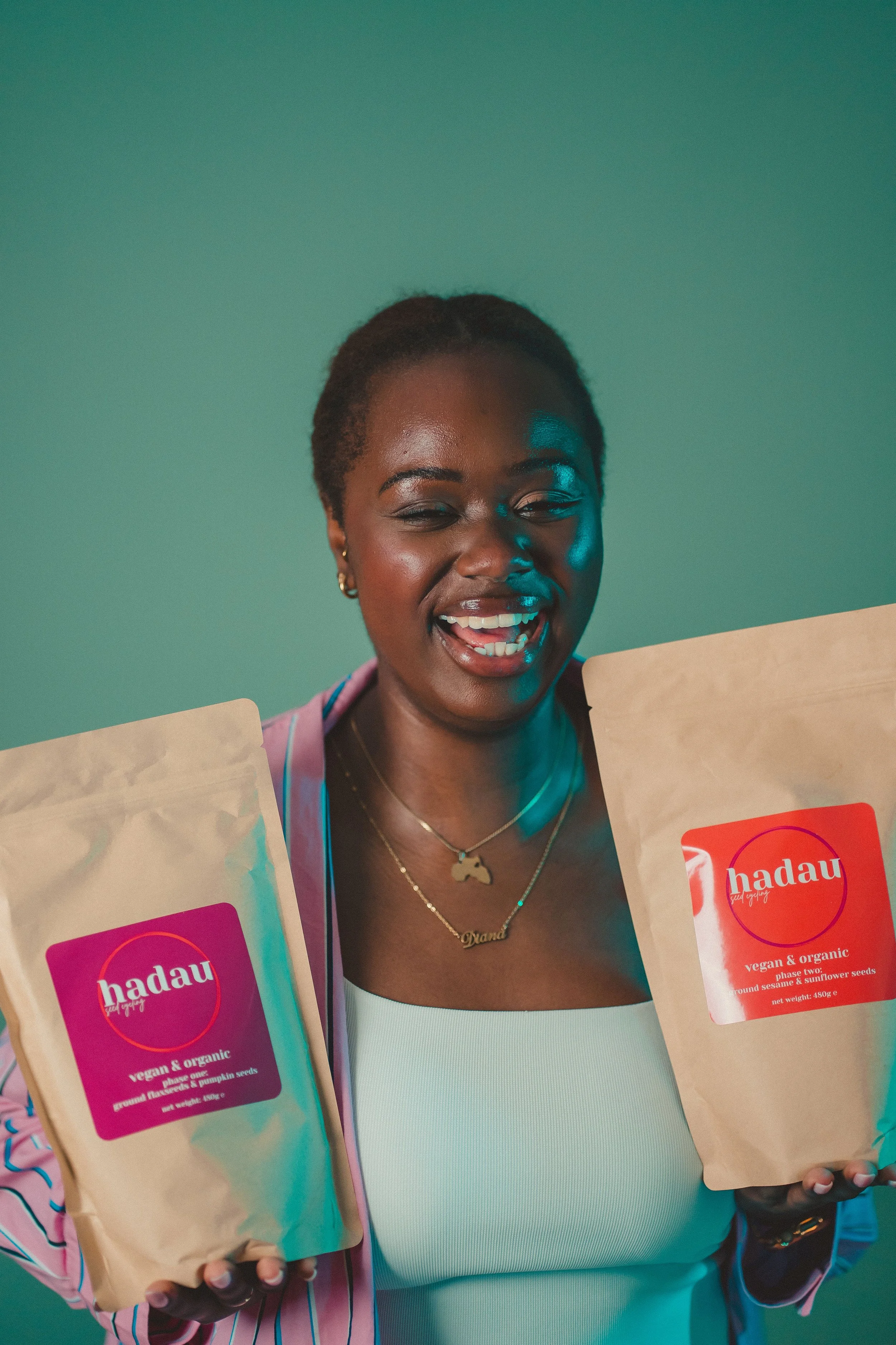A smiling woman holding two packages of Hadau vegan and organic ground sesame and sunflower seeds, standing against a green background.