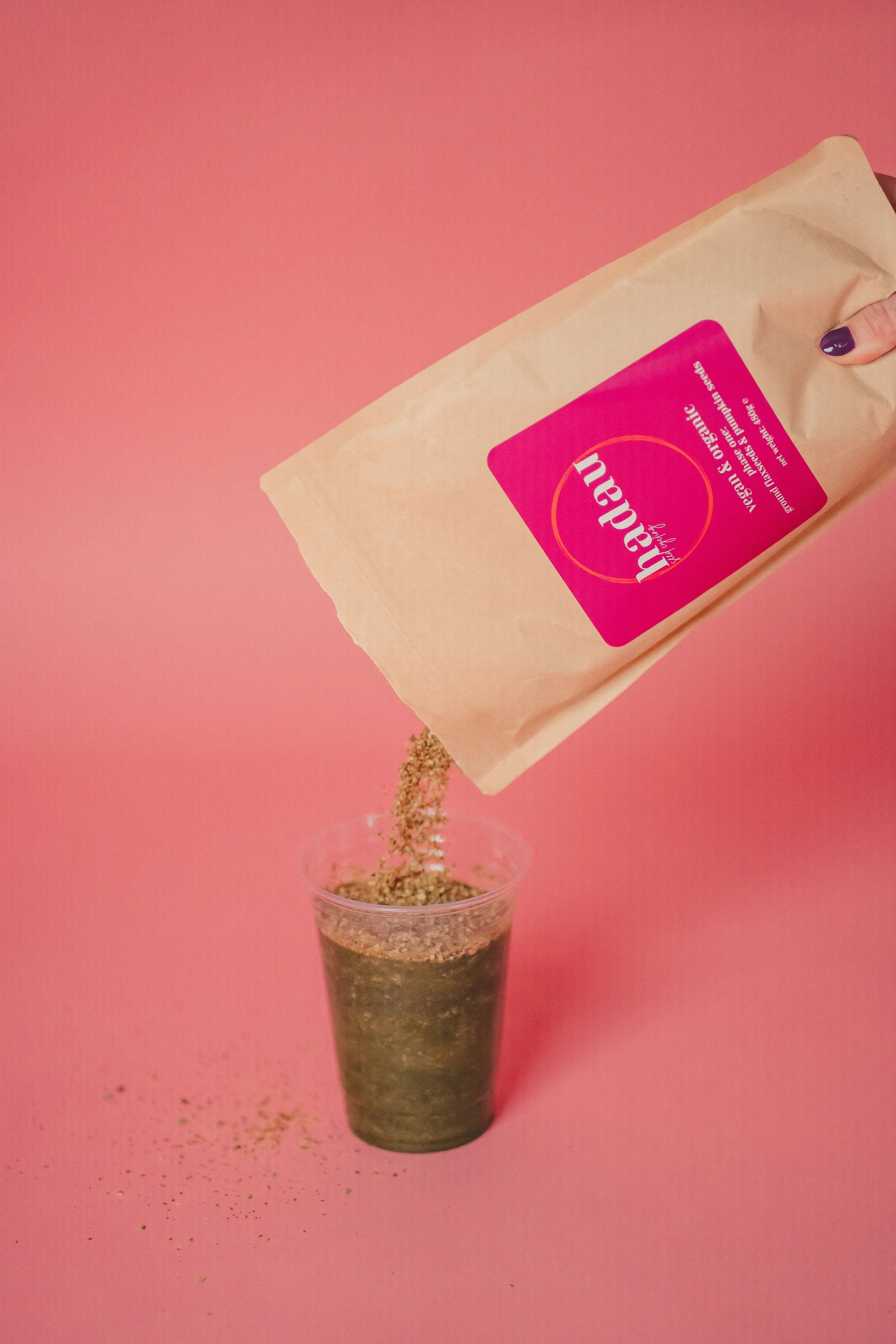 Hand pouring powdered matcha tea from a pink labeled paper bag into a clear glass on a pink background.