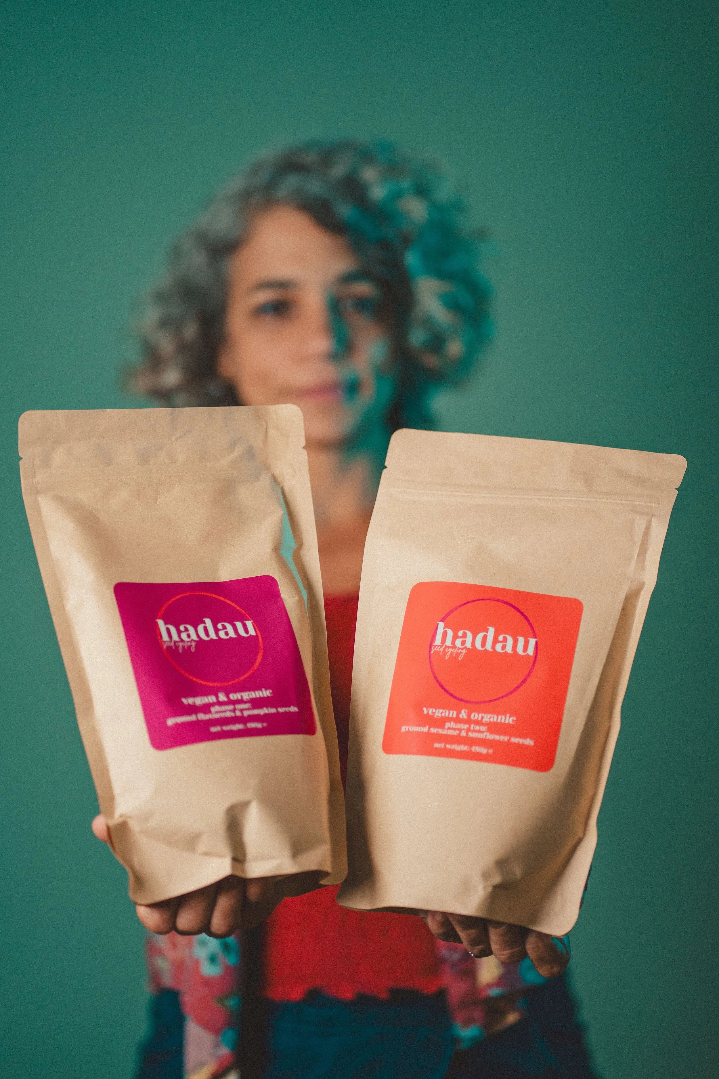 A woman with curly hair holding two bags of vegan organic sesame and sunflower seeds, with a blurred green background.