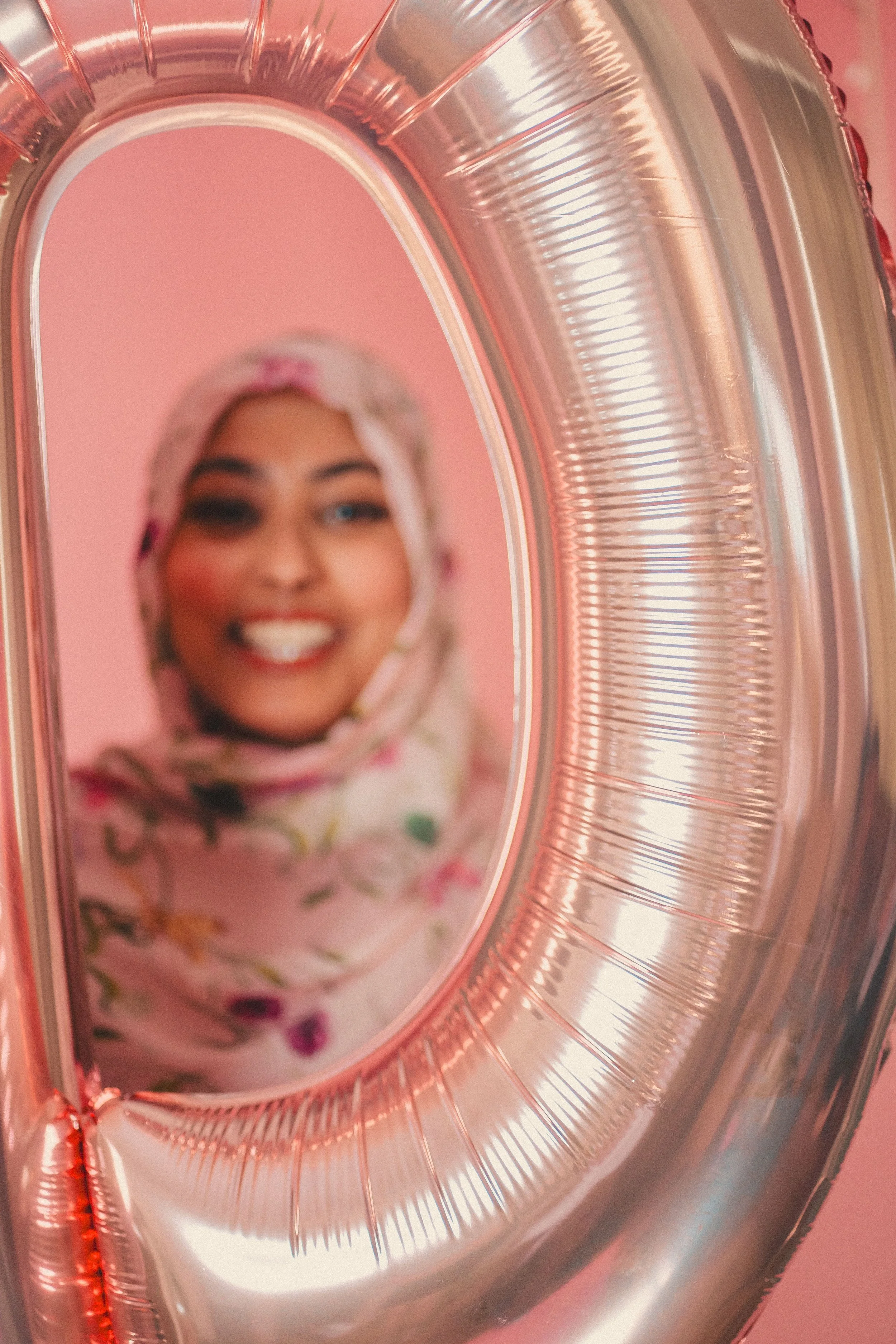 A smiling woman wearing a floral hijab looking into a mirror shaped like a rose gold balloon.