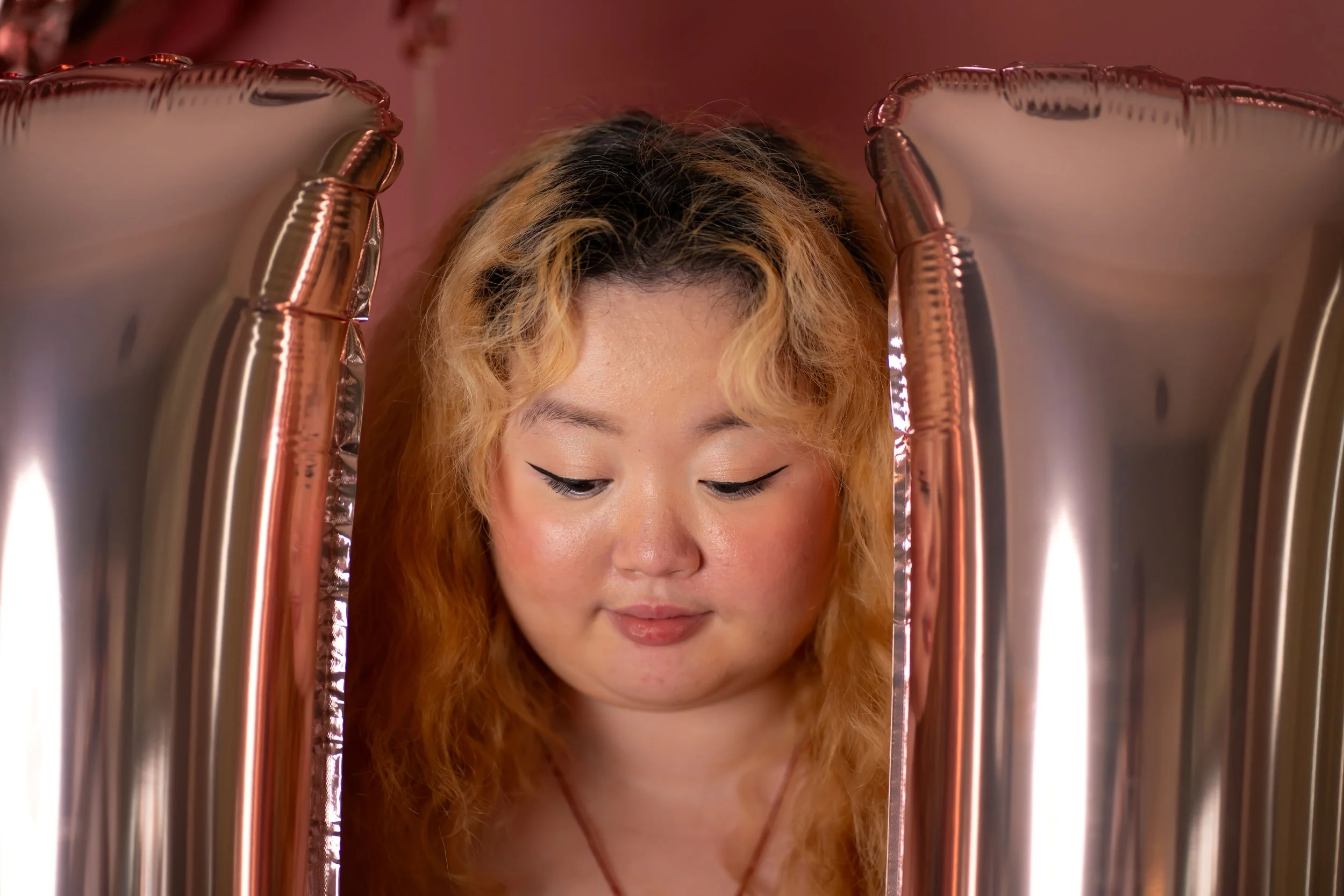 A young woman with curly blonde hair and winged eyeliner looking down, surrounded by metallic pink balloons.