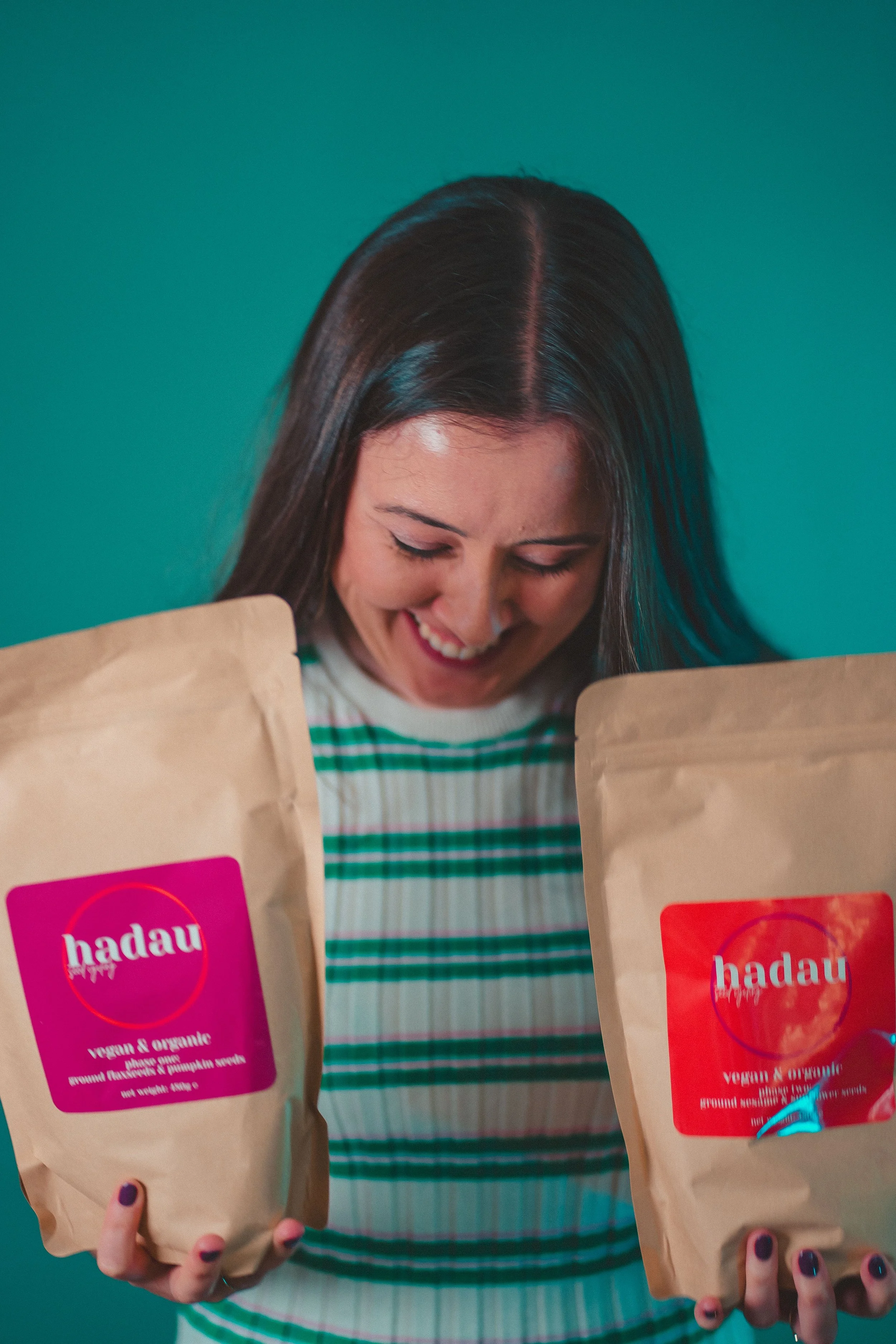 A woman holding two brown paper bags with pink and red labels that read 'hadau' and 'vegan & organic,' smiling and looking down.