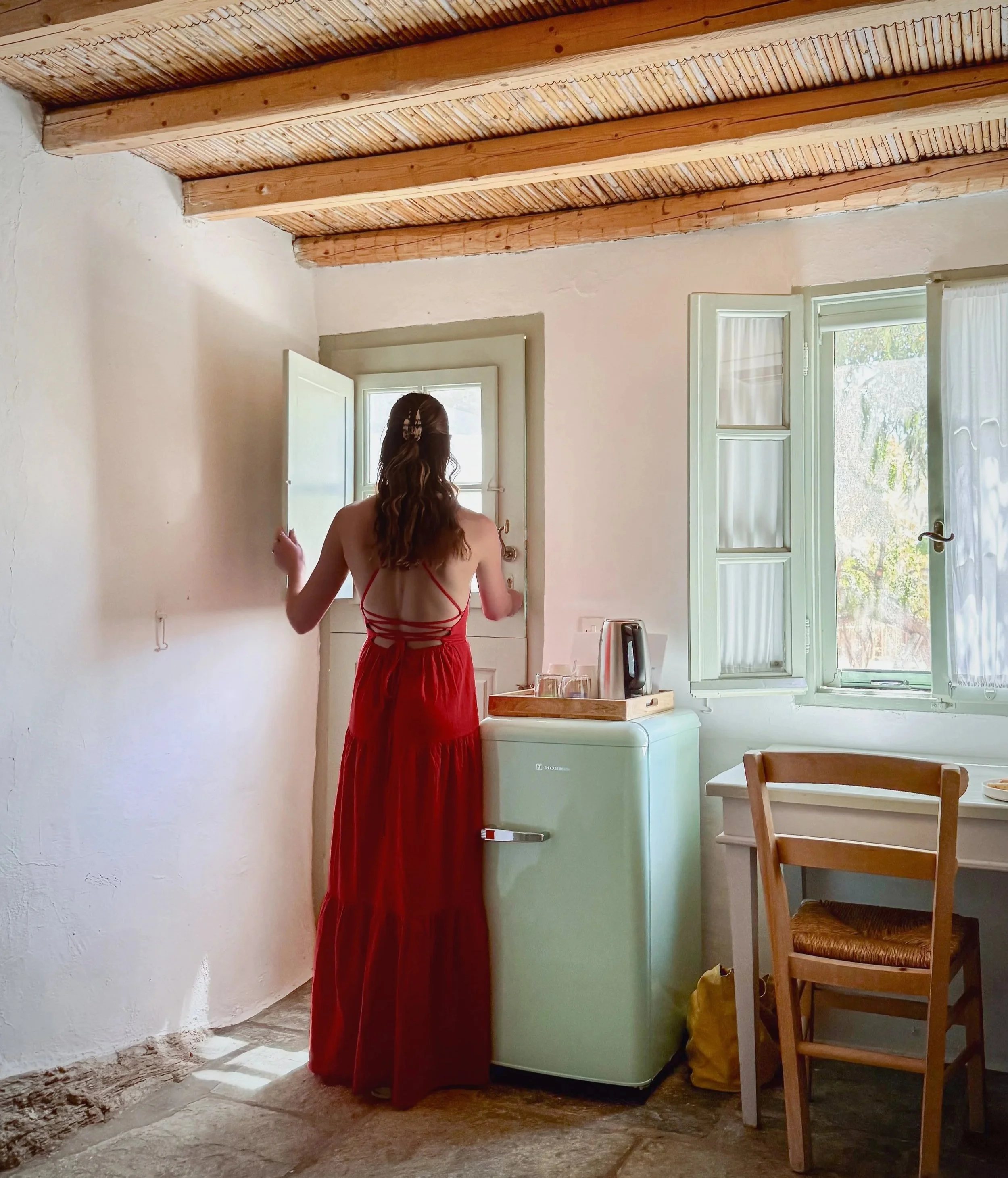A woman in a red dress standing in a bright, rustic room of Nature Hotel Eco Living in Serifos, Greece, with a vintage refrigerator, white walls, wooden ceiling beams, and open windows.