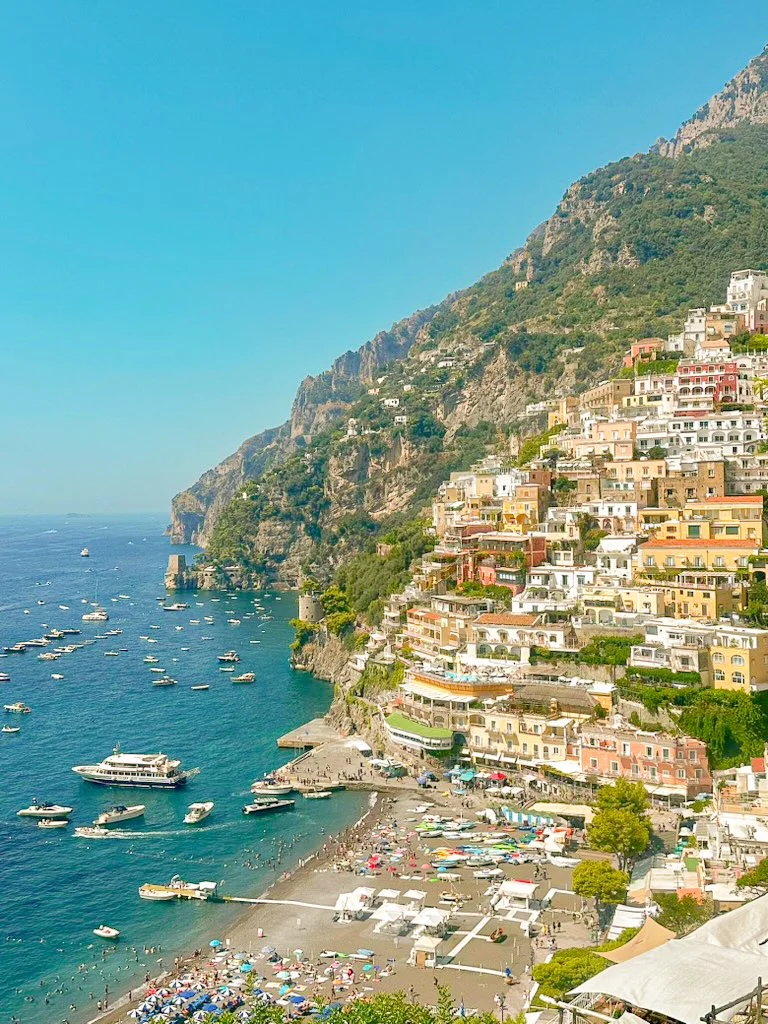 Colorful buildings stacked on the Amalfi coast line overlooking a the beach with umbrellas and boats in the water, in Positano Italy.