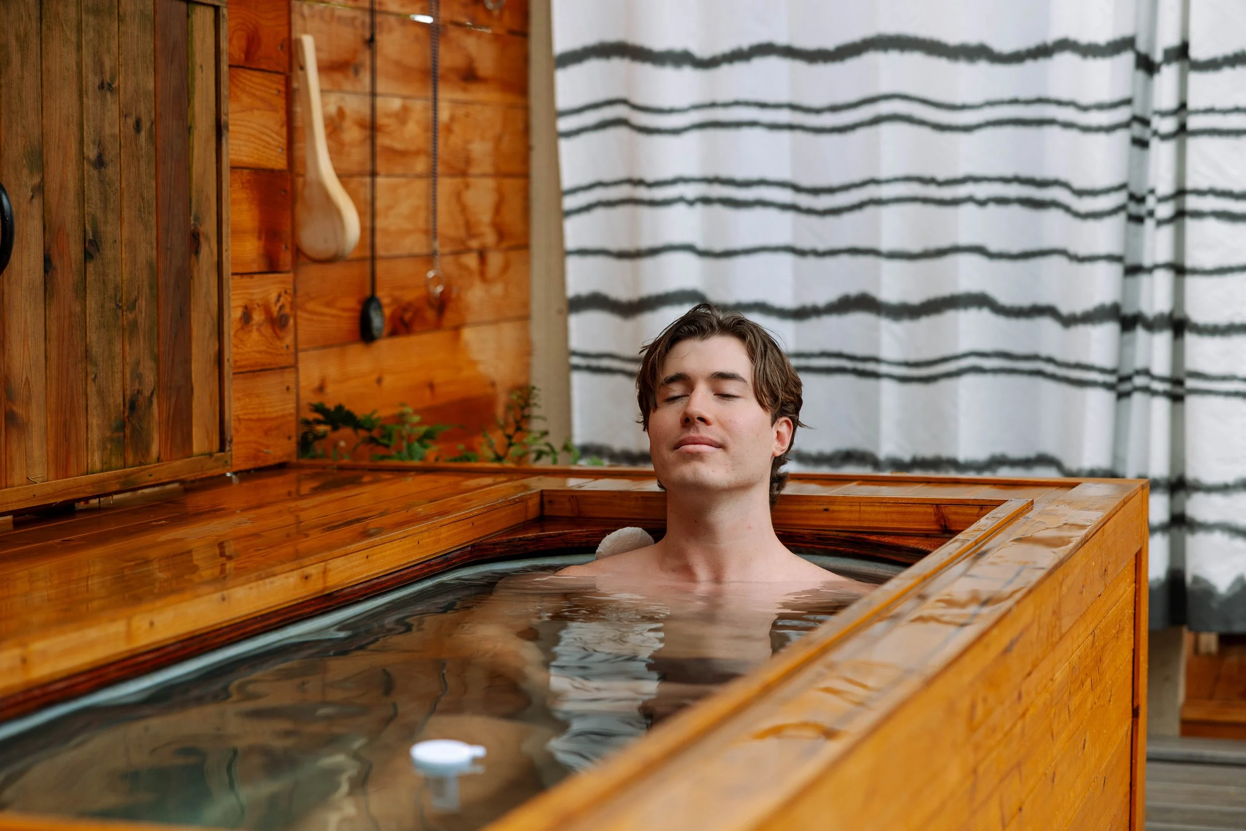 A man relaxing in a wooden hot tub with his eyes closed indoors.