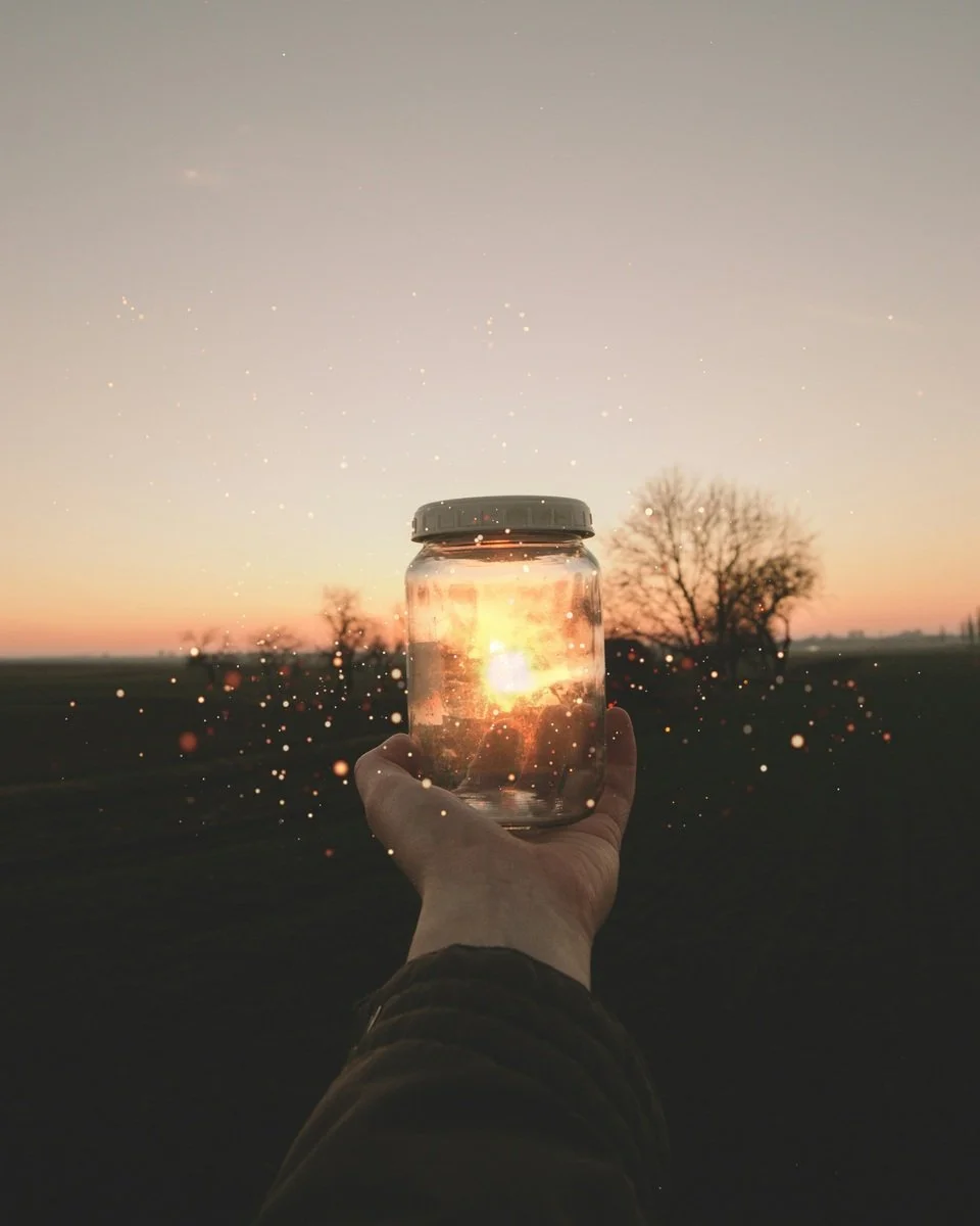 Person holding a jar with the setting sun inside, against a sunset sky and silhouette trees in the background.