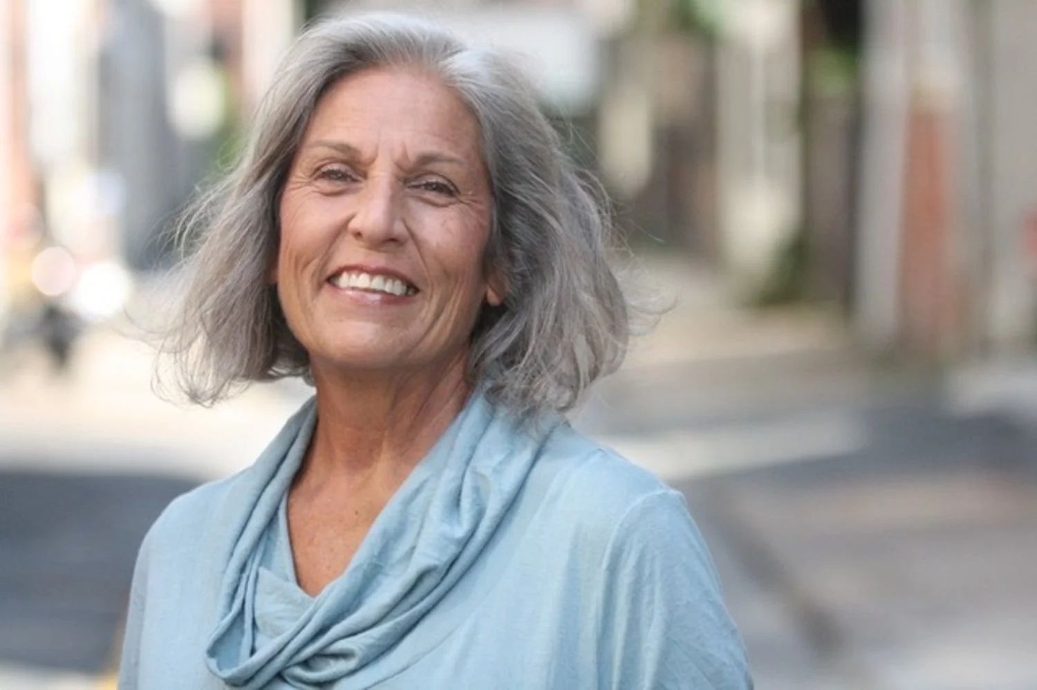 A smiling elderly woman with gray hair standing outdoors in an urban setting, wearing a light blue shirt and a matching scarf.