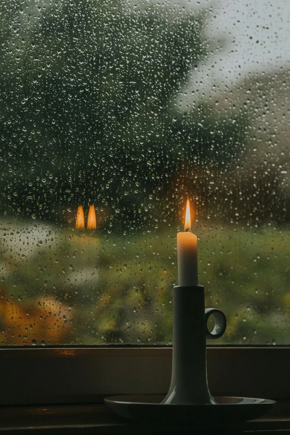 A lit candle on a white holder sitting on a windowsill with raindrops on the windowpane, blurred trees and grass visible outside on a rainy day.
