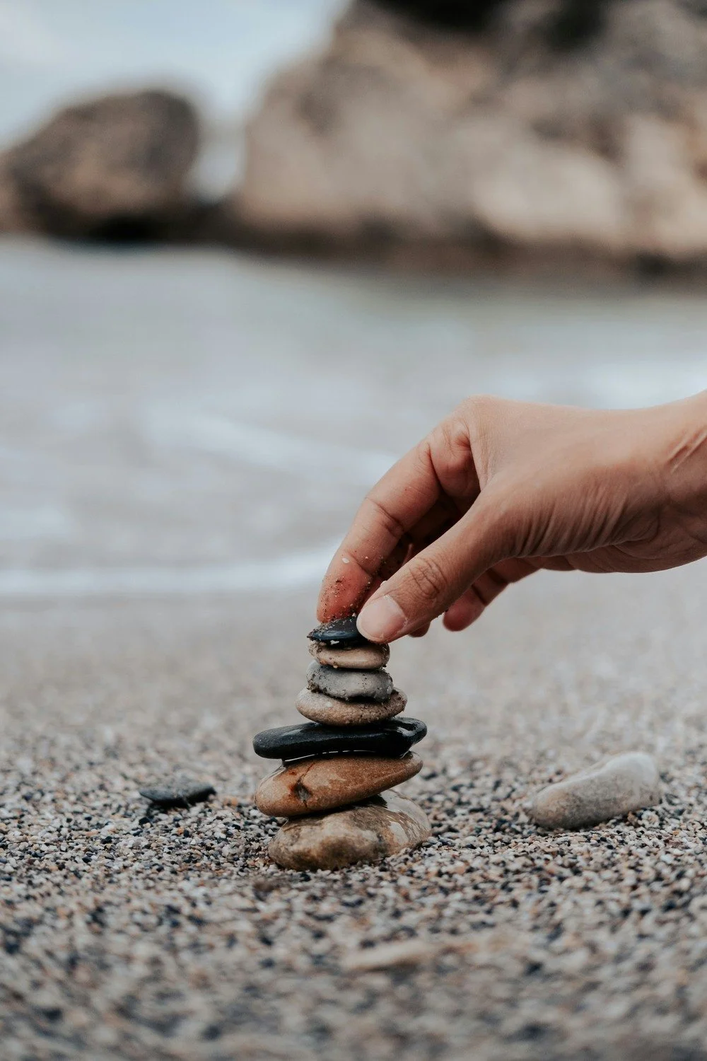 A hand stacking small, flat stones on a sandy beach with the ocean and larger rocks in the background.