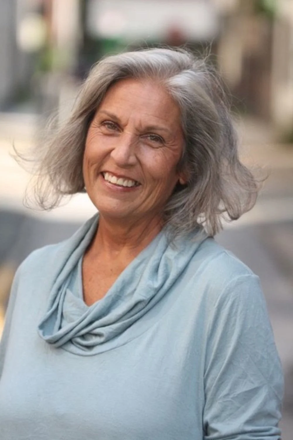 An older woman with gray hair smiling outdoors in a light blue top with a matching scarf.