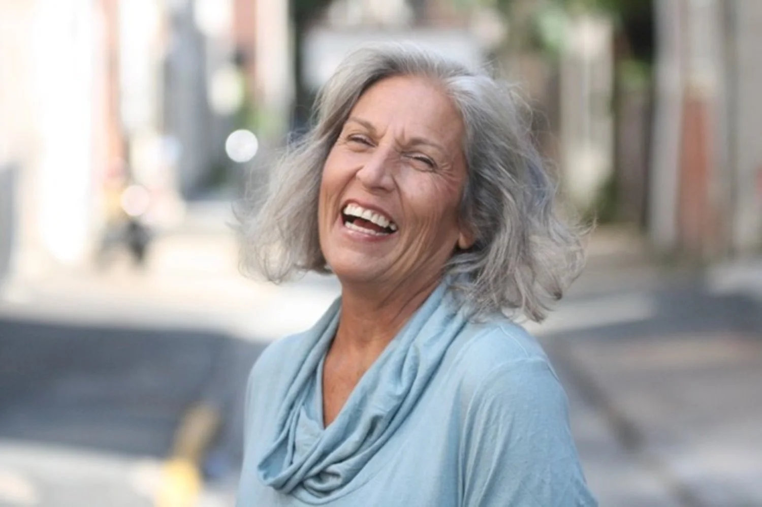 An elderly woman with gray hair laughing outdoors in a city street.