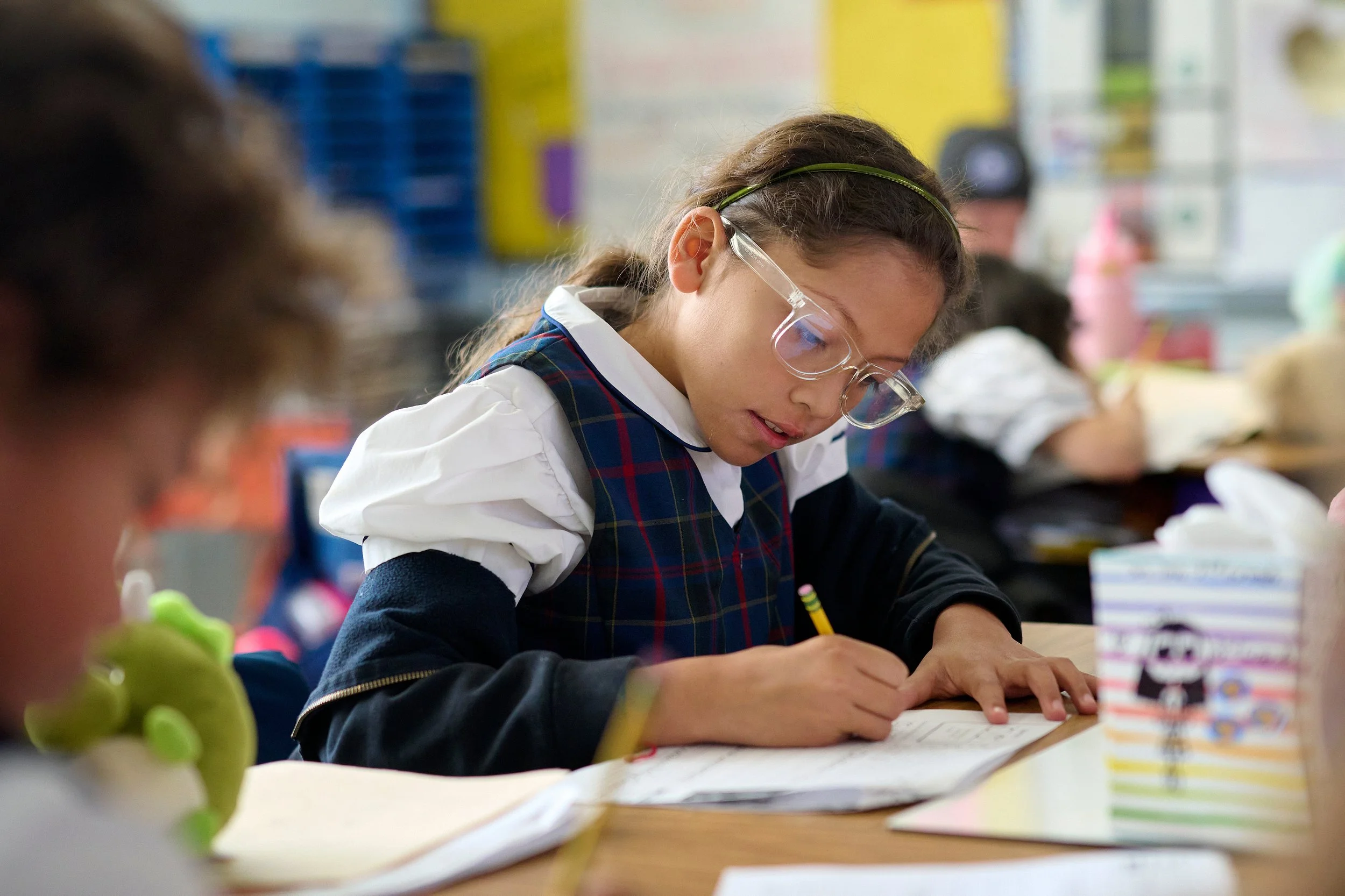 Girl in school uniform writing in notebook at desk in classroom.