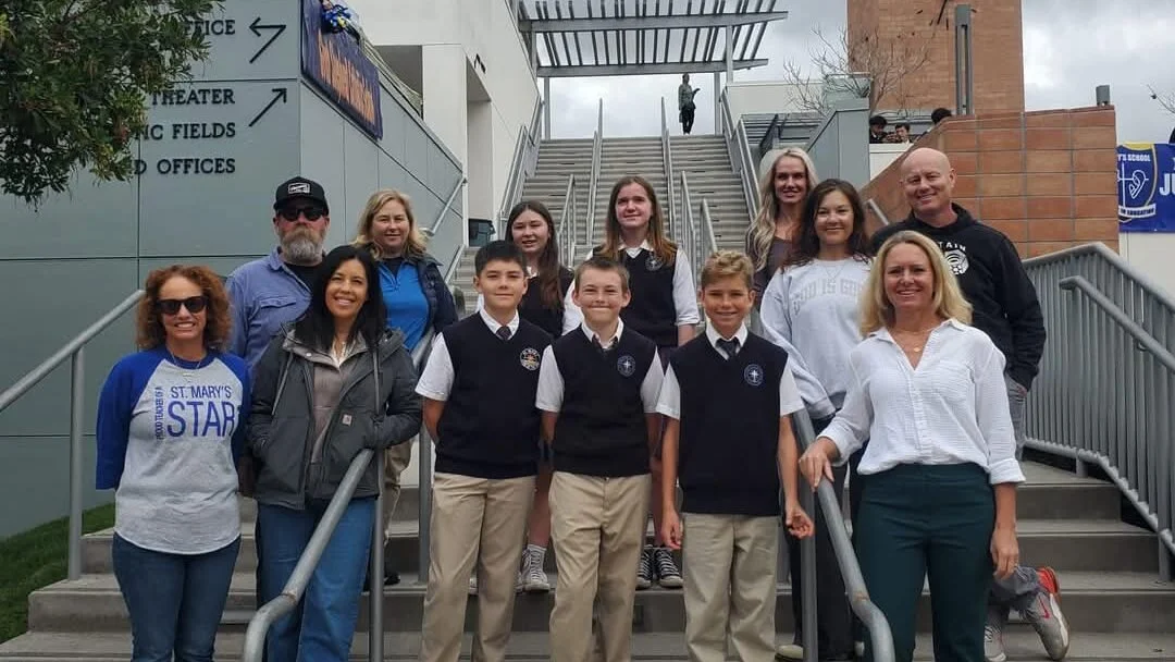 A group of students and adults posing on outdoor stairs in front of a school, with some wearing uniforms and others in casual clothing.