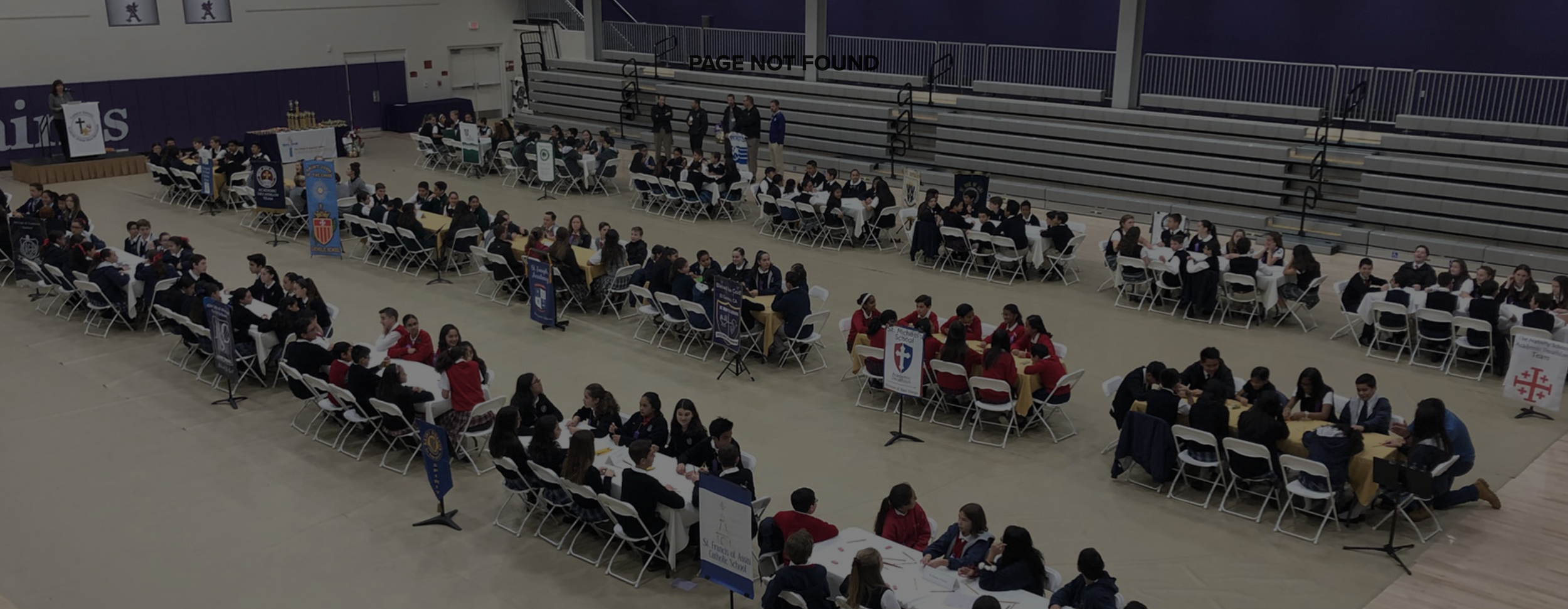 Students at tables participating in a chess tournament inside a gymnasium with banners and a speaker on the left.