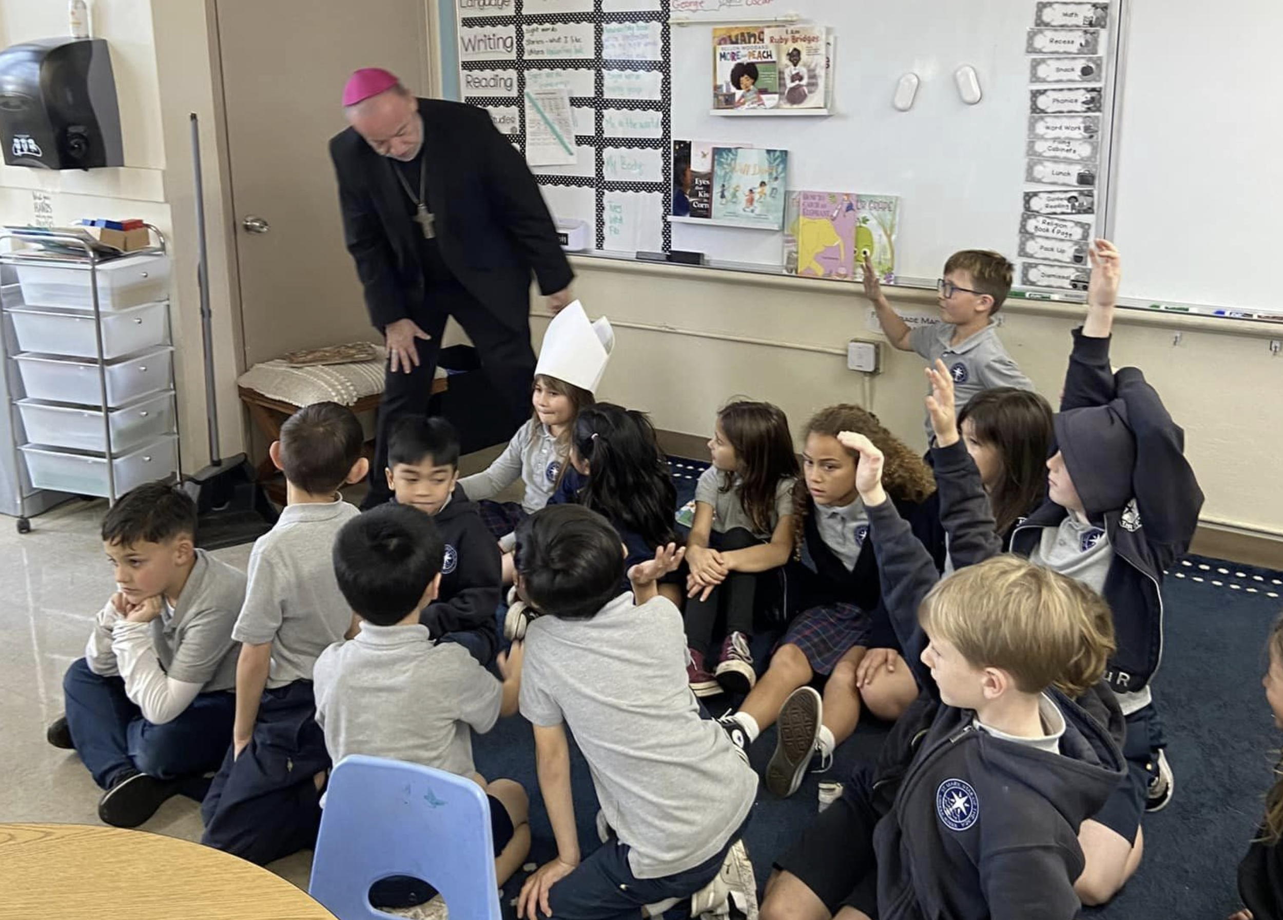 A classroom scene with a group of elementary school students sitting on the floor, some raising their hands, and a man dressed as a bishop with a pink skullcap standing nearby, engaging with the students.