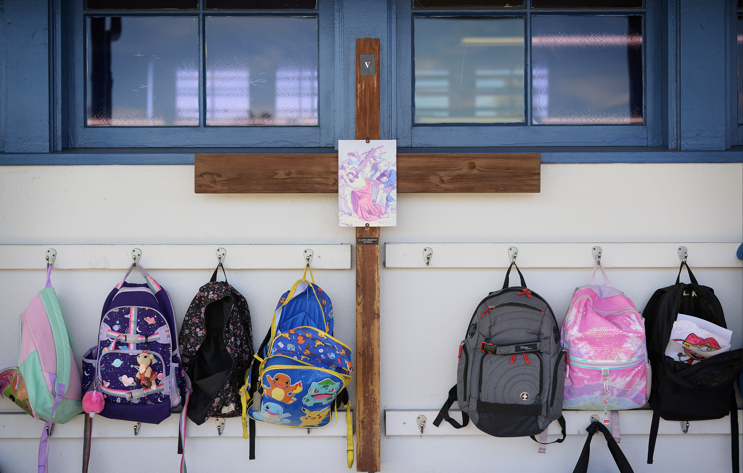 School backpacks hanging on hooks outside a classroom, with a wooden cross and a picture of Jesus Christ in front of a window.